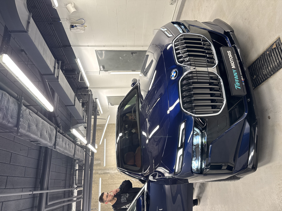 A dark blue BMW SUV is the central subject, positioned indoors within a brightly lit facility, likely a professional garage, detailing studio, or showroom in Warsaw, Poland. The image is presented vertically, showing the car from a high-angle frontal perspective.
The car appears meticulously clean and polished, with sharp reflections of the numerous overhead fluorescent strip lights visible on its dark paintwork. Its distinctive large kidney grille, sleek LED headlights, and the classic BMW emblem are clearly visible. A temporary license plate holder is affixed to the front, prominently displaying "TIFFANY MOTORS" in white text on a black background, with "DYNAMIC MOTORS GROUP" and a small BMW M logo underneath.
To the left of the car, a man with short dark hair, glasses, and a beard stands next to the open driver's side door. He is wearing a dark hoodie and appears to be observing or interacting with the vehicle, with his right hand raised towards his face in a thoughtful gesture.
The environment is characterized by an industrial aesthetic, featuring exposed grey ventilation ducts running across the ceiling, numerous bright fluorescent tube lights, and a grey, possibly brick or textured, wall. A floor drain grate is visible on the right side of the car, suggesting a workshop or wash bay area. The bright artificial lighting indicates that the time of day is not relevant to the scene. The overall impression is one of professional presentation or inspection of a high-end vehicle.