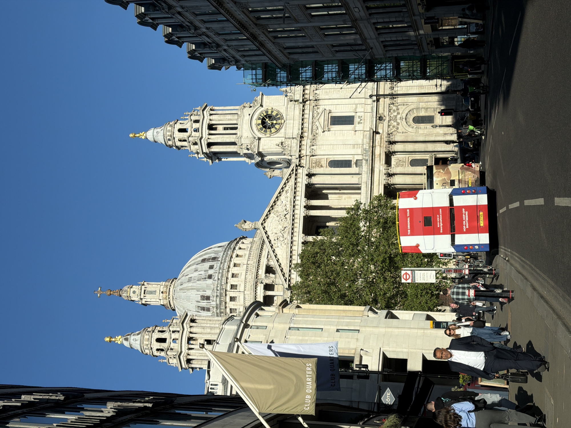 The image presents a bright, sunny daytime scene in London, United Kingdom, centered around St. Paul's Cathedral. The majestic white-stone cathedral, with its iconic dome, intricate Baroque architecture, and visible clock face on one of its towers, dominates the background against a clear blue sky. Scaffolding is visible on a building adjacent to the cathedral.
In the midground and foreground, a typical London street scene unfolds. A red, white, and blue double-decker "THE ORIGINAL TOUR" sightseeing bus is prominently featured on the right side of the street, displaying "HOP-ON, HOP-OFF SIGHTSEEING" and the website "theoriginaltour.com", along with the license plate "YJII TVO". Several pedestrians are visible on the sidewalk, including a man in a dark suit standing near the foreground, and others congregating near a bus stop marked by a London Underground roundel symbol. Motorcycles and other vehicles are also present on the street.
The clear sky and strong shadows indicate bright, sunny weather, likely late morning or early afternoon. Two flags displaying "CLUB QUARTERS" are visible in the lower left, associated with a building also showing a "CÔTE" sign. The overall scene captures the bustling activity around a major London landmark.