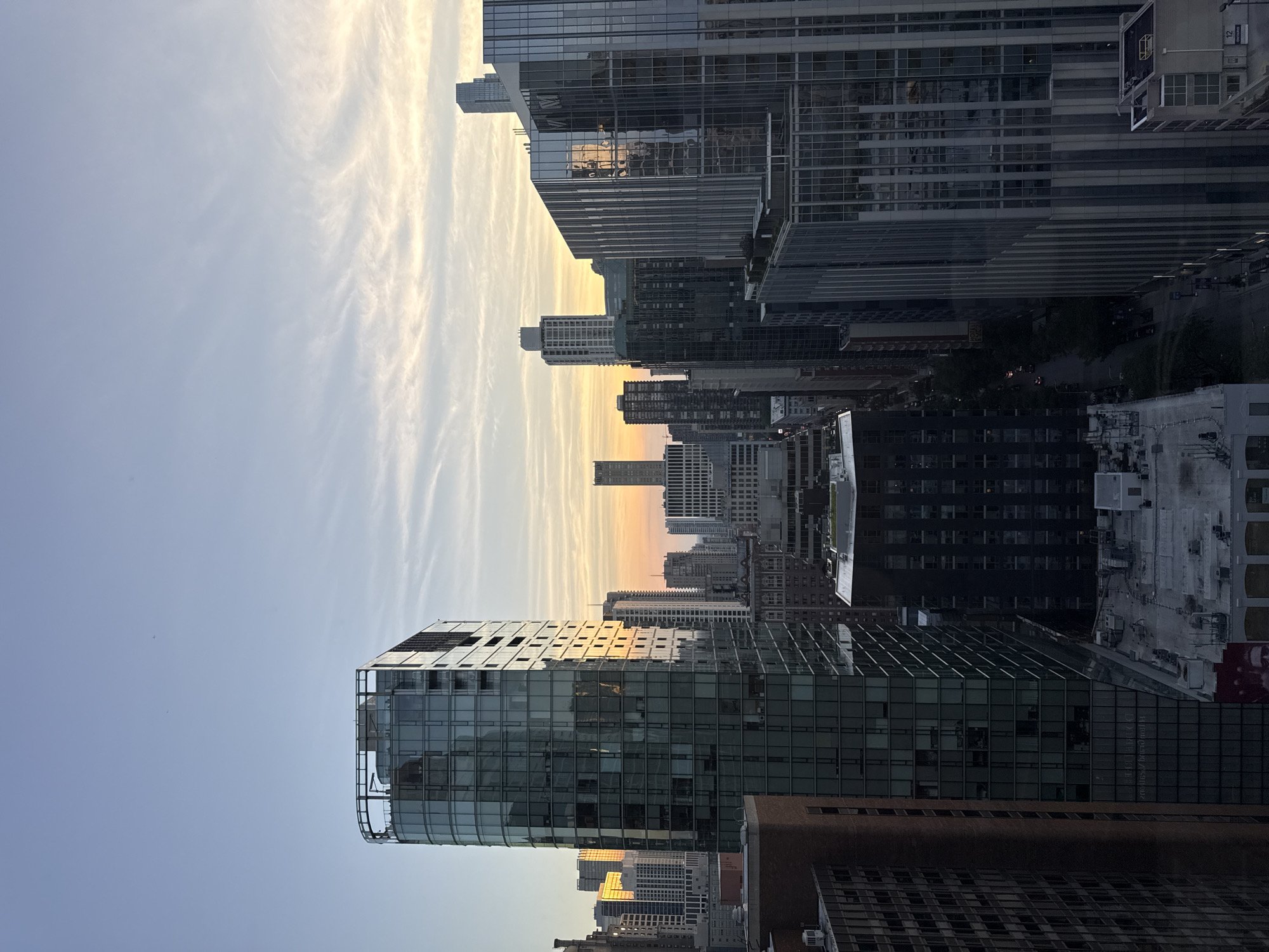 This is an aerial, full shot of a cityscape in Chicago, USA, during the golden hour. The sky is a pale blue with wispy white clouds, transitioning to a soft yellow and orange gradient near the horizon. The city is densely packed with skyscrapers of varying heights and architectural styles, many of which are glass-fronted and reflect the sky and surrounding buildings. The most prominent building in the foreground is a tall, curved skyscraper with a glass facade and a visible rooftop structure. Other buildings are a mix of modern high-rises and older, more traditional brick buildings. The street level is not clearly visible but hints of trees and streetlights can be seen between the buildings. The image appears to be taken from a high vantage point, possibly another building or a drone.