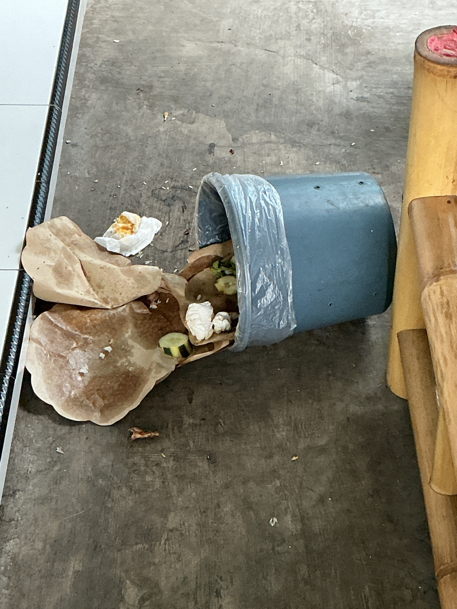 A tipped-over blue trash can lies on a concrete floor, spilling its contents of food waste and brown paper wrappers. Some of the discarded food items include pieces of cucumber and cauliflower. The scene is located outdoors, with a vertical white and metal structure on the left and a portion of a wooden structure on the right. The location is specified as Magelang, Indonesia.
