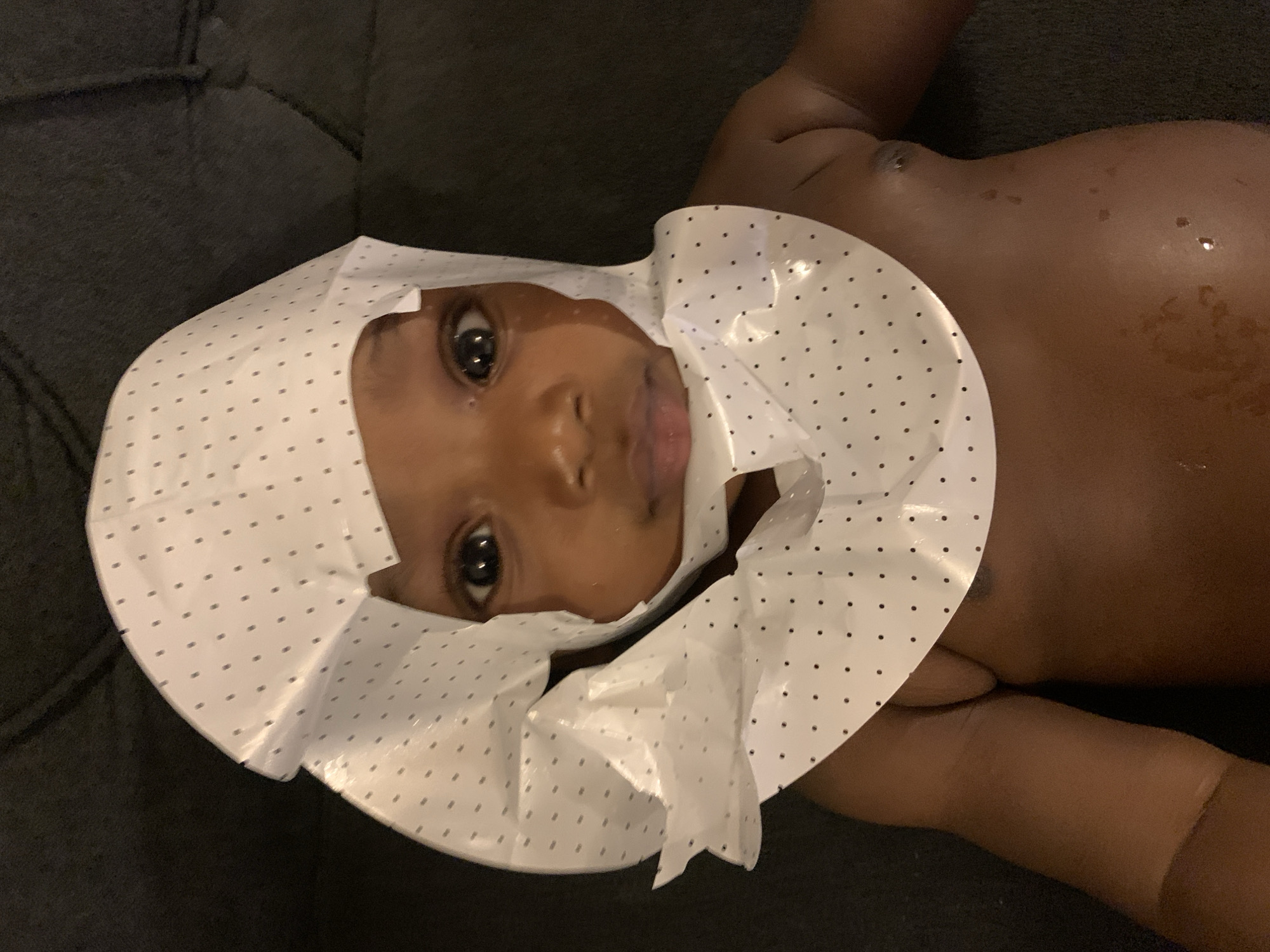 The image shows a baby lying on a dark gray surface, possibly a couch. The baby is wearing a white shower cap with black polka dots. The baby is dark-skinned and appears to be wet. The baby's eyes are open and looking at the camera. There is no indication of the location being Barakin Akawo, Nigeria, or any other specific location.