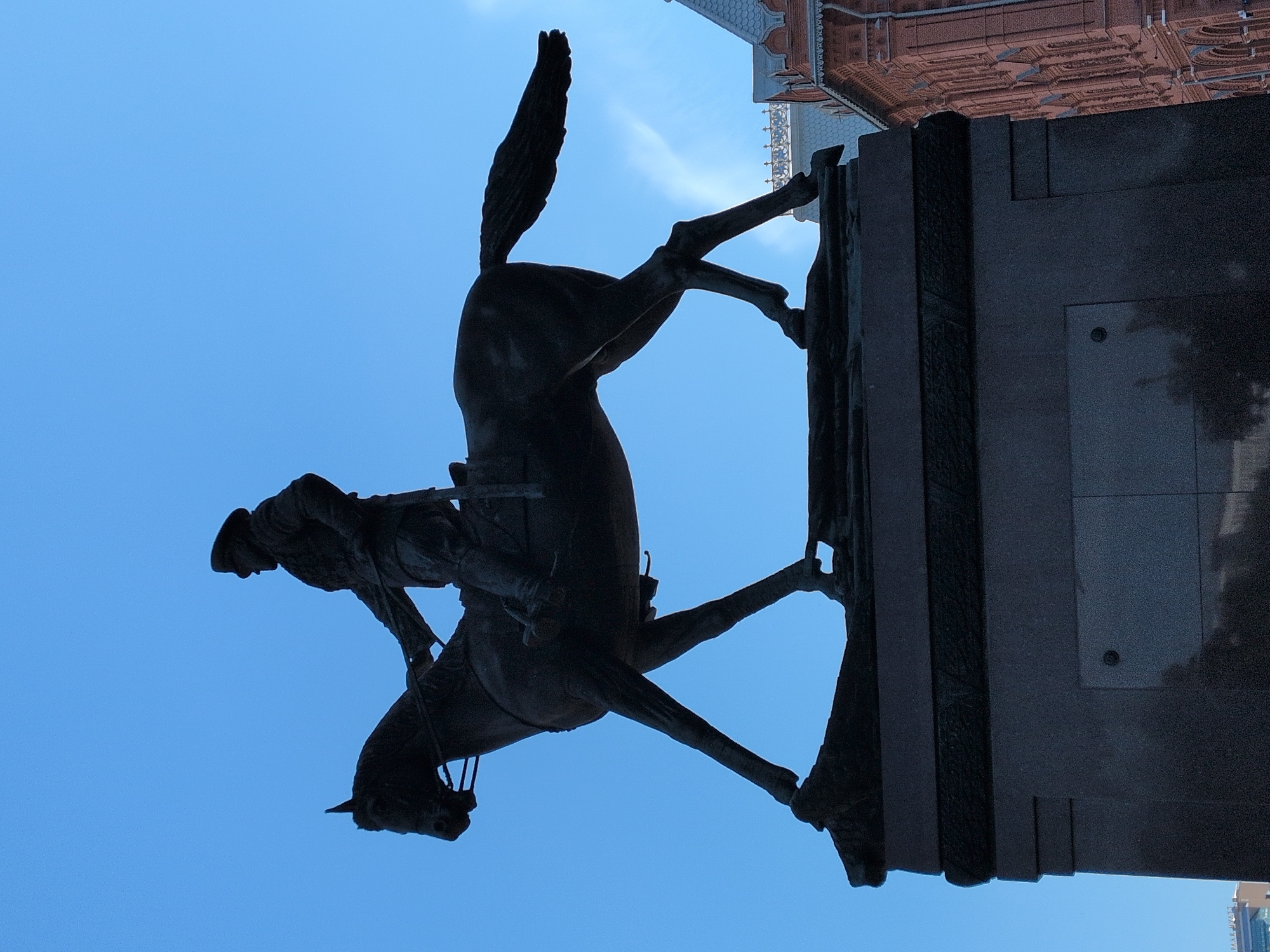 An equestrian statue is prominently featured, captured from a low-angle perspective against a clear, bright blue sky. The statue, appearing as a dark silhouette, depicts a male figure astride a horse that is rearing on its hind legs. The horse's front legs are lifted high, and its tail is angled upwards, conveying a sense of motion and power. The rider is seated firmly in the saddle.
The statue is mounted on a large, multi-tiered pedestal or base made of dark stone, which occupies the right side of the frame. Partially visible behind the statue and pedestal is a building with distinctive architectural elements. This building appears to be constructed of red or terracotta-colored brickwork and features ornate decorative details along its roofline and upper sections, possibly indicating a historical or elaborately designed structure.
The strong silhouette of the statue and pedestal suggests that the photograph was taken during the day with the sun positioned behind the subject, casting it in shadow. The weather appears clear and sunny. No discernable text is visible on the statue, pedestal, or building.
