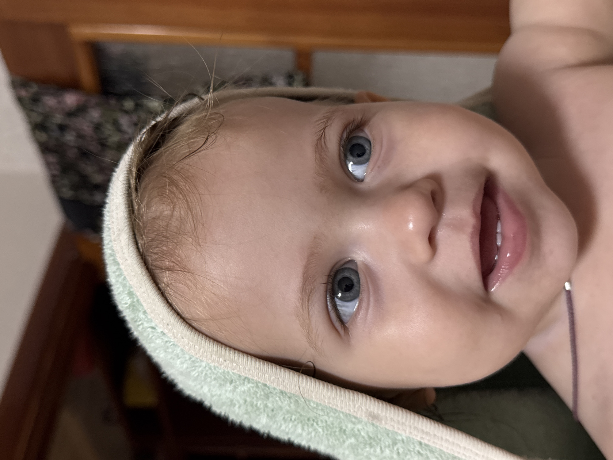 This is a close-up, horizontally oriented, slightly angled photograph of a young child, likely a baby or toddler, wrapped in a light green towel. The child has bright blue eyes that are looking directly at the camera with a curious and engaging expression. Their mouth is slightly open, revealing a hint of teeth and glossy lips. Fine, light-colored hair is visible around the child's forehead and temples, with a few strands sticking out. The towel has a textured edge and appears soft.
The background is out of focus but suggests an indoor setting. There are hints of wooden furniture, possibly a bed frame or dresser, and a patterned fabric in darker colors. The lighting appears to be artificial and soft, creating a warm and intimate atmosphere. The overall emotion conveyed is one of innocence and playful curiosity. No visible text is present in the image. The location context of Chaikovsky, Russia, does not provide specific visual cues within this image.