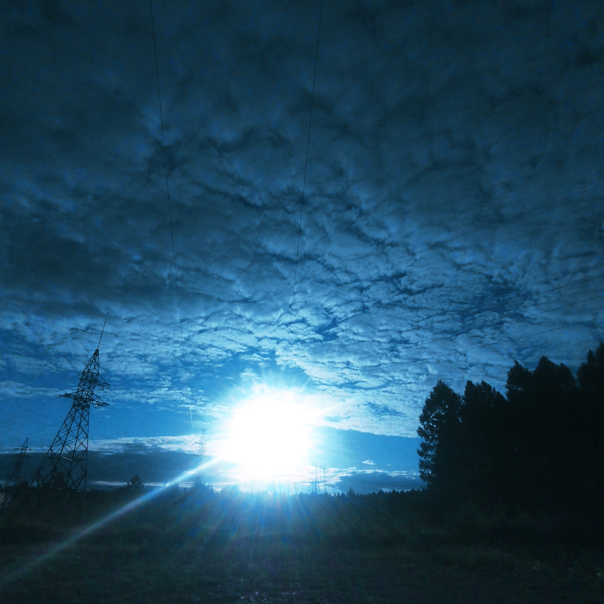 The image depicts a landscape scene under a heavily clouded, blue sky, illuminated by the bright sun. In the foreground, a silhouetted treeline runs along the bottom right of the frame. To the left, a tall, lattice-style electrical pylon stands prominently. Further in the background, other electrical pylons are visible, some appearing smaller and more distant. The sun is a brilliant, blown-out white light in the upper center of the image, casting lens flares and light rays across the scene. The ground appears to be a grassy field, also silhouetted in dark tones. The overall impression is of an industrial landscape with natural elements, possibly taken during sunrise or sunset due to the low angle of the sun and dramatic lighting. The location is identified as Pokosnoye, Russia.