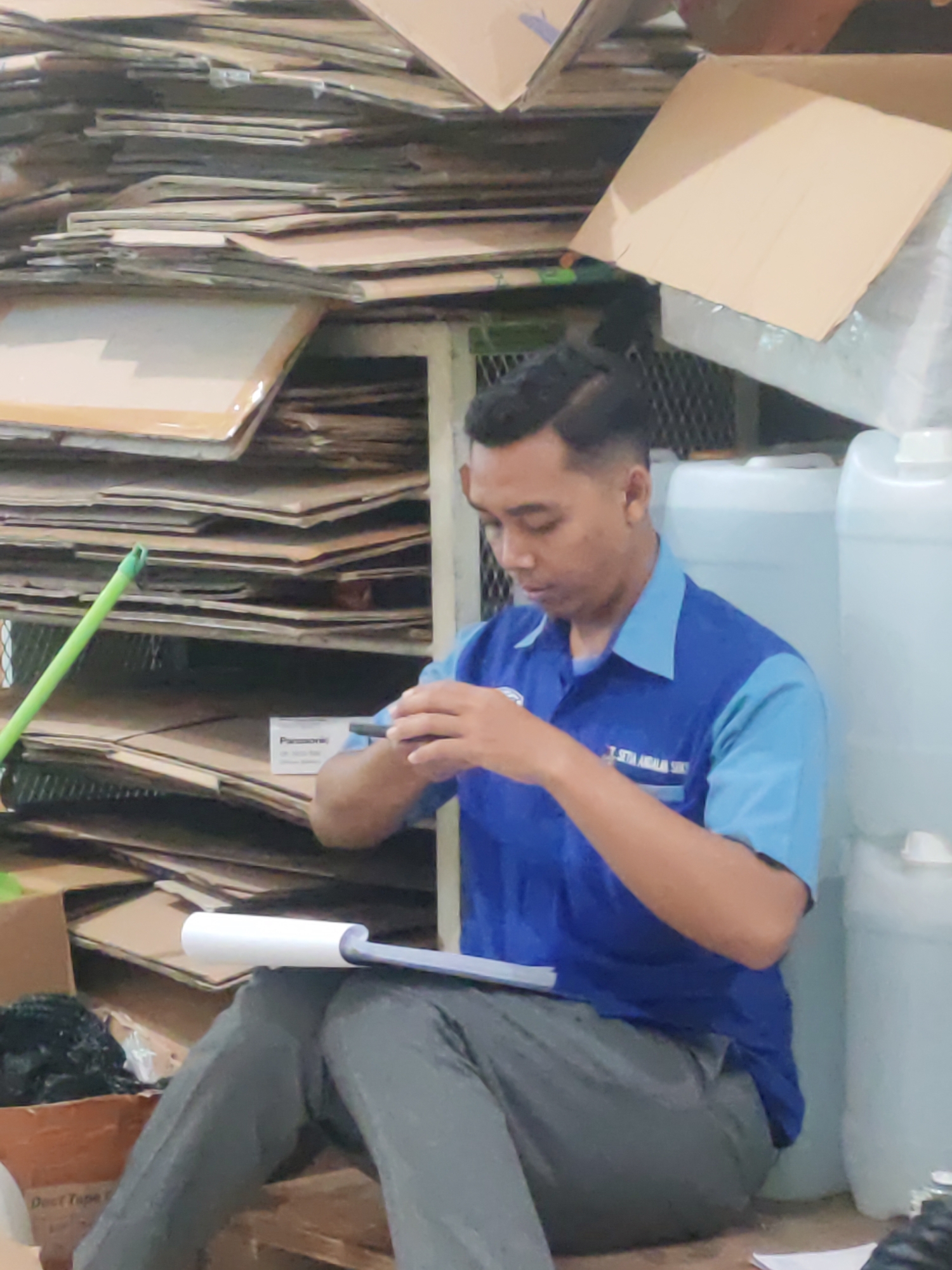 A young man wearing a blue and light blue polo shirt and grey pants is sitting on a wooden pallet in what appears to be a storage area. He is holding a black stylus in his right hand and looking down at a tablet, which rests on his lap. A white roller-like object is placed on top of the tablet. In the background, there are stacks of cardboard boxes and large white plastic containers. To the left, a green broom is leaning against a shelf. The overall environment suggests a warehouse or a backroom of a business. The man is likely an employee at this location in Pepelegi, Indonesia.