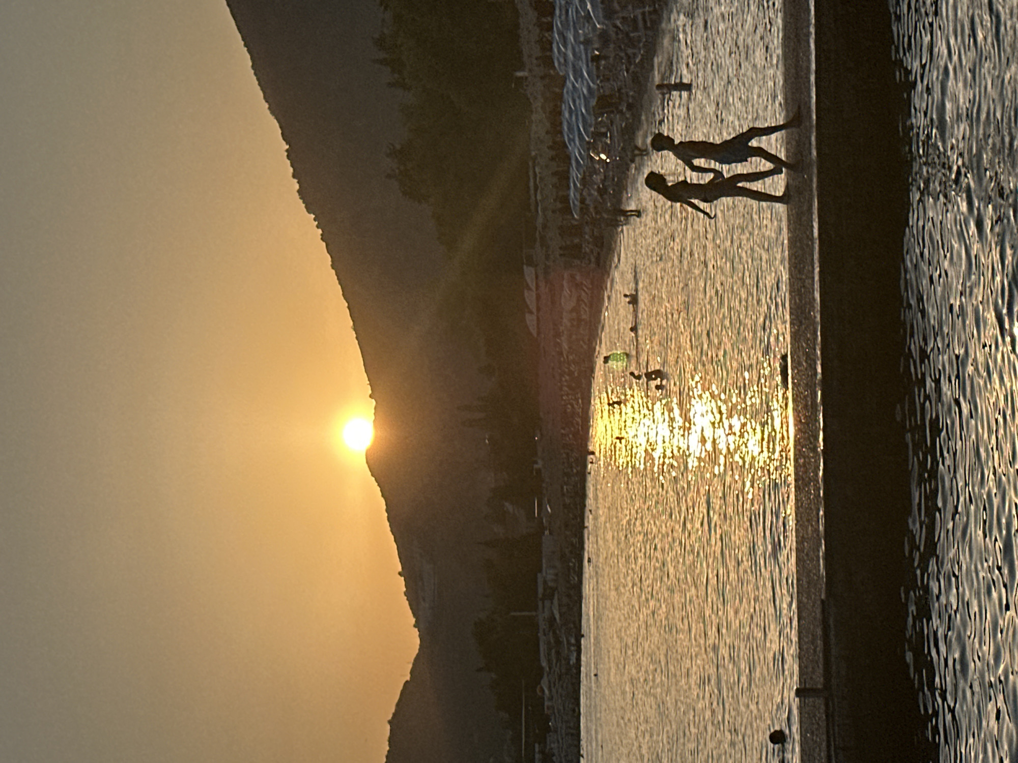 The image depicts a sunset at a beach in Budva, Montenegro. The sun is setting behind a mountainous silhouette, casting a golden glow over the water and sky. The sea is relatively calm, with gentle waves reflecting the sunlight. In the foreground, a dark, flat structure, possibly a pier or breakwater, extends into the water. Two silhouetted figures, a man and a woman, are walking together on this structure.
In the mid-ground, a beach is visible, lined with numerous beach umbrellas, indicating a populated area. Several people are in the water, some swimming and others standing near the shore. The water closest to the beach is shallow and filled with more sunbathers and swimmers.
In the background, the mountains are dark silhouettes against the bright, orange-yellow sky, with the sun's rays extending outwards. The overall scene captures a tranquil evening at a popular coastal resort.