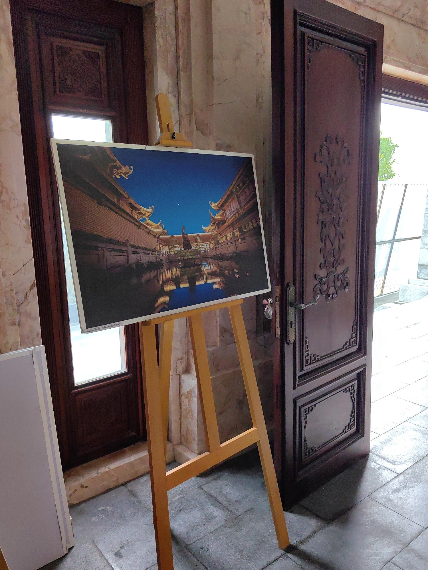 A photograph shows an easel standing on a tile floor, displaying a framed image of a building in Hong Gai Ward, Vietnam. The building is ornate and has a body of water in front of it. The scene is lit by a clear, dark blue sky, with stars visible above the structure.
Behind the easel and the framed image are two open, carved wooden doors. Beyond the right-hand door, the outside world is partially visible. To the left of the easel is a white rectangular object.
The flooring is composed of grey tiles. The walls in the scene are marbled with pink and beige tones.