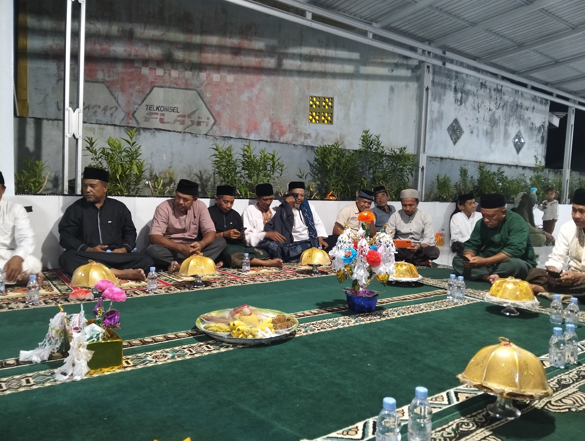 The image depicts a gathering of men, likely for a religious or cultural event, seated on a green carpet in what appears to be an outdoor or semi-outdoor setting. The background shows a wall with faded graffiti and some foliage. The men are dressed in traditional attire, including caps and long-sleeved shirts. Some are looking down at their phones, while others are engaged in conversation. In the foreground, there are several food items displayed on platters, as well as decorative items like golden domes and floral arrangements. Bottles of water are also scattered throughout the scene. The overall impression is one of a communal meal or observance taking place in a relaxed, informal atmosphere. The presence of the "TELKOMSEL FLASH" graffiti on the wall suggests the location might be in Indonesia, and specifically the city of Petapa, based on the provided information.