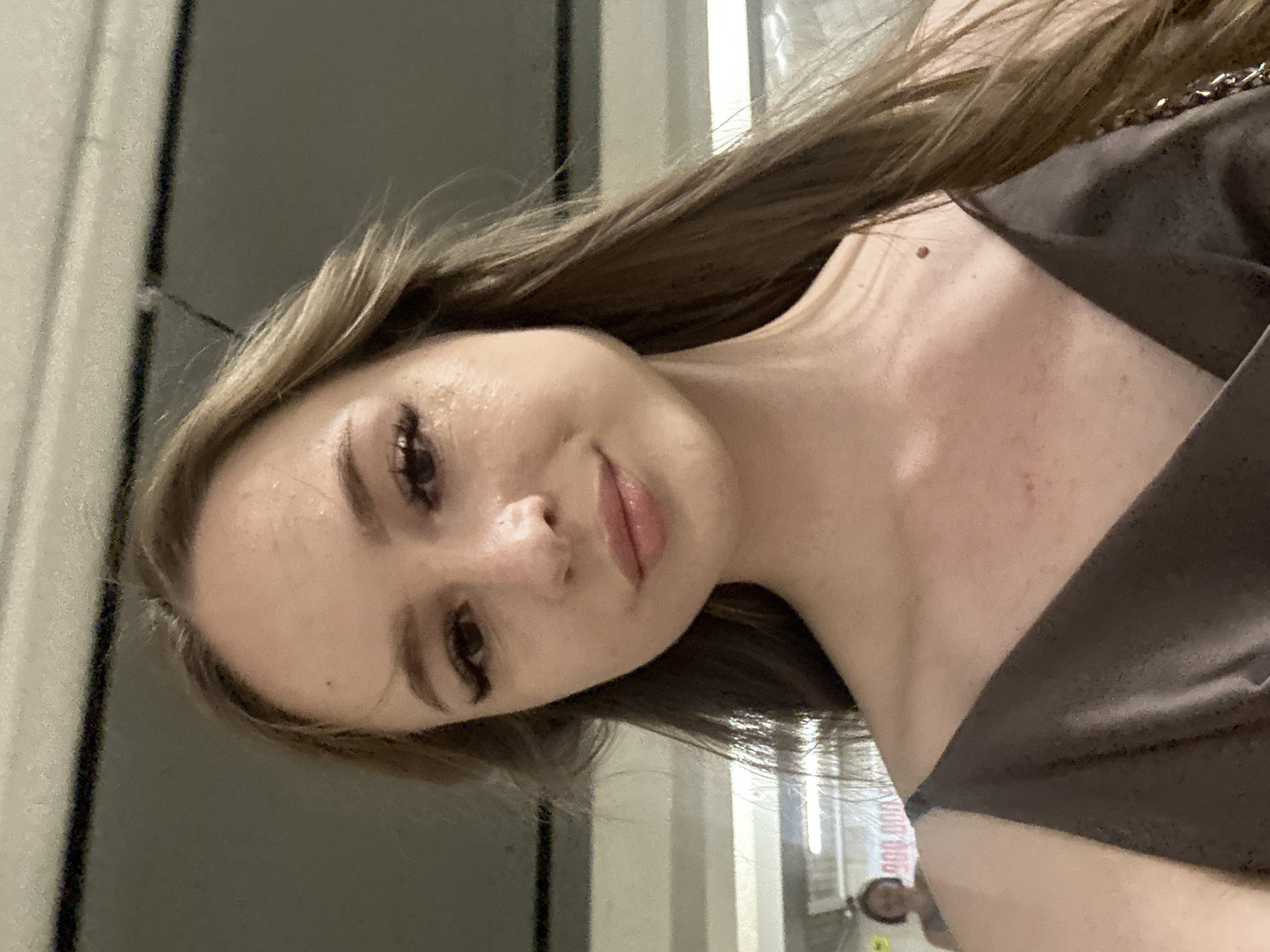 A young Caucasian woman with long brown hair and light skin is pictured in a close-up, self-taken photograph. She is wearing a dark brown dress with thin straps. Her makeup includes winged eyeliner and mascara. In the background, a blurry figure of another person is visible in what appears to be an indoor setting with fluorescent lighting. The location is identified as Unknown, Unknown.