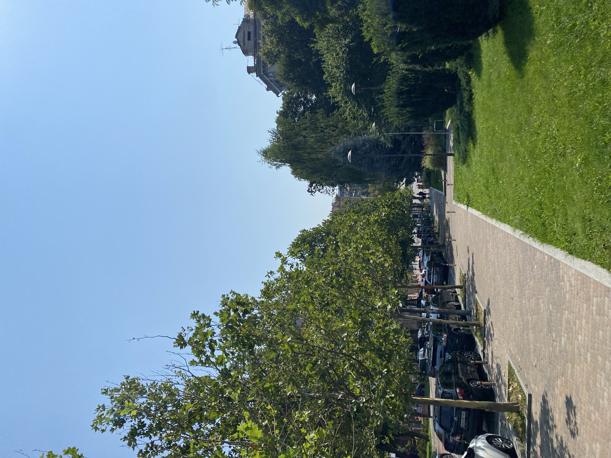 The image depicts a street scene in a city under a clear blue sky. On the left side of the frame, a row of parked cars lines the street, mostly dark-colored sedans and SUVs. The cars are parked alongside a paved walkway made of brick-like pavers, which is bordered by a strip of grass and trees. The trees are deciduous, with lush green foliage, and are planted at regular intervals along the street, casting dappled shadows on the walkway.
Beyond the parked cars, a building with a mansard roof and visible antennas can be seen in the distance. The right side of the image features a manicured lawn with neatly trimmed bushes and several lampposts. A few people are walking on the walkway in the background, appearing as small, distant figures. The overall impression is of a well-maintained urban environment on a sunny day. The license plate of one car is visible and reads "KA 6787 PE".