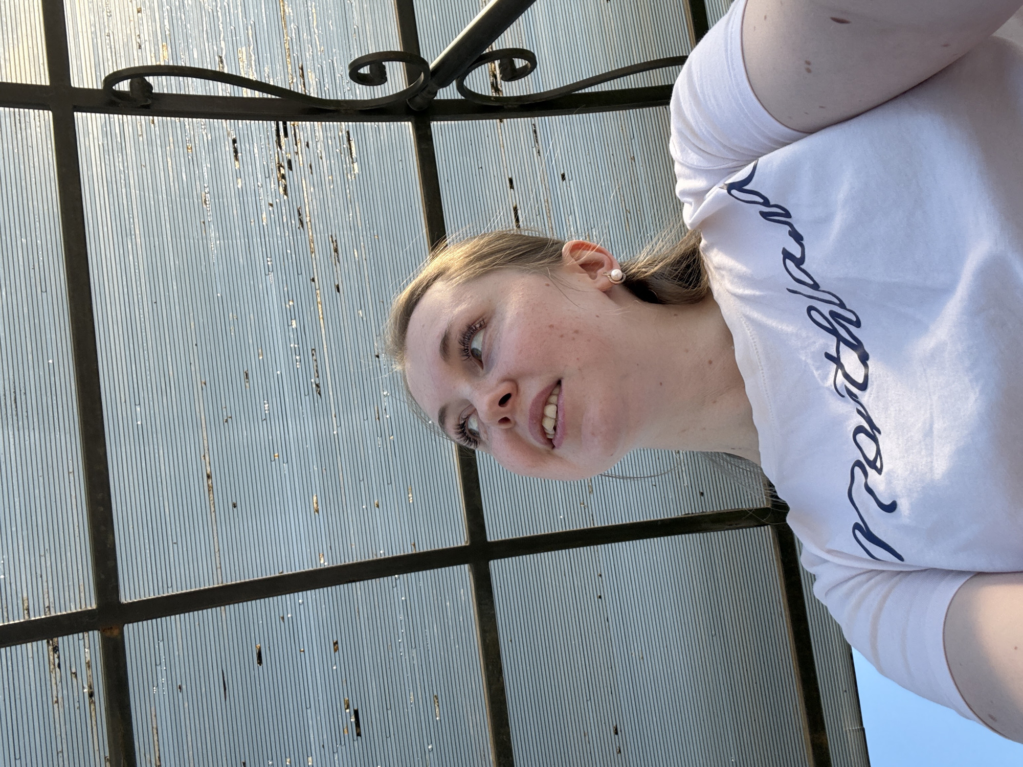 A low-angle, medium shot depicts a woman in a white t-shirt with stylized lettering, positioned in front of a gridded metal and translucent panel structure. The woman is fair-skinned, with light brown hair partially visible near her face and ear, and is looking upward with a slight smile. The background consists of a geometric pattern created by the metal grid against a translucent material, possibly a roof or overhead structure. The location is unknown, and the city is also unknown.
