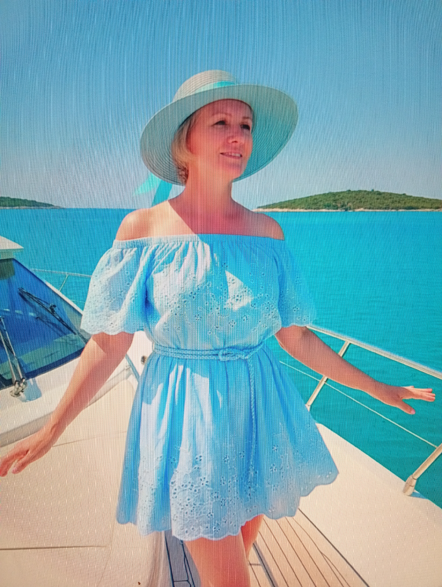 A light-skinned woman with blonde hair, wearing a light blue off-the-shoulder dress and a light blue wide-brimmed hat, stands on the deck of a white boat. The woman is facing to her right, with her arms outstretched. She appears to be smiling and looking out at the ocean. The background is a bright blue sky and turquoise water, with a distant green island. The deck of the boat is visible in the foreground, with a white railing. The lighting is bright and sunny, suggesting it is daytime. The image appears to be taken from a slightly low angle, looking up at the woman.
