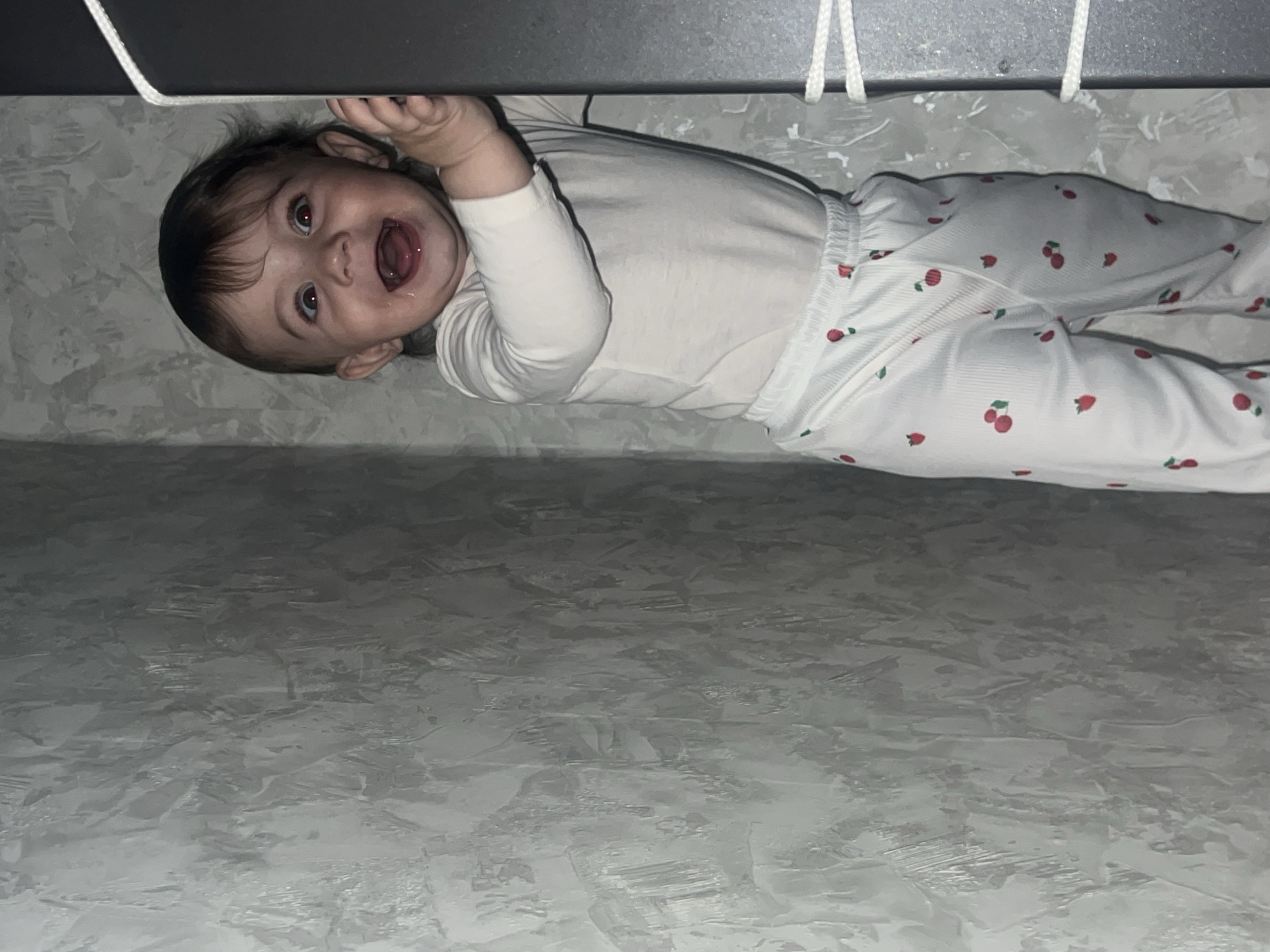 A baby lies on a surface with a textured, grayish-white appearance. The baby is wearing a long-sleeved white shirt and patterned pants with red cherry motifs. The baby's head is tilted towards the viewer, and its mouth is open, showing teeth. The baby is reaching up, possibly touching something out of frame. A section of what appears to be a gray edge or possibly a bar and two white strings are visible in the upper left corner. The context of the city of Elena, Bulgaria, is not evident from this image.