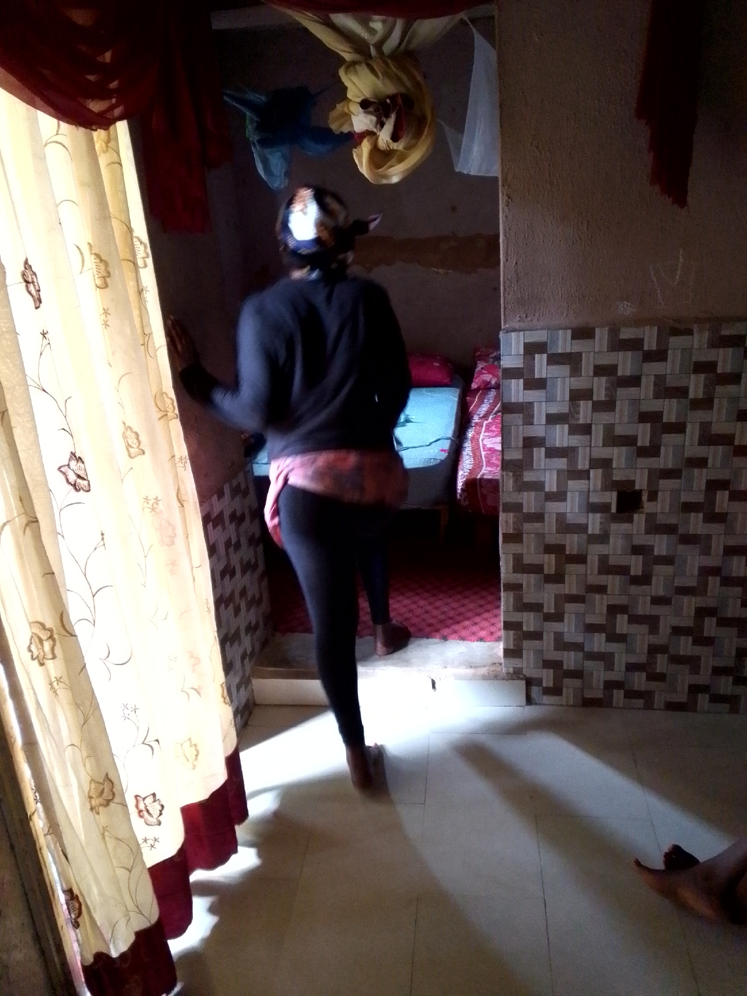 The image depicts an indoor scene, possibly in Jos, Nigeria. A person, viewed from behind and slightly to the side, is standing in a room. They are wearing a dark long-sleeved top, a pink patterned garment around their waist, and black leggings. A patterned headscarf covers their head. The person is standing near a doorway or opening, with their right hand resting on a wall or partition.
To the left of the person, a cream-colored curtain with floral patterns and a dark red trim hangs, partially obscuring the view. Sunlight streams in from the left, casting shadows on the tiled floor.
In the background, a bed with blue and red bedding is visible, suggesting a bedroom or sleeping area. The walls have a textured finish, with a tiled section on the right featuring a geometric pattern of brown and beige squares. Draped fabric, in shades of red and yellow, hangs from the ceiling in the upper part of the frame, adding to the cluttered appearance of the environment. Another person's legs are visible in the lower right corner of the image, lying on the floor.