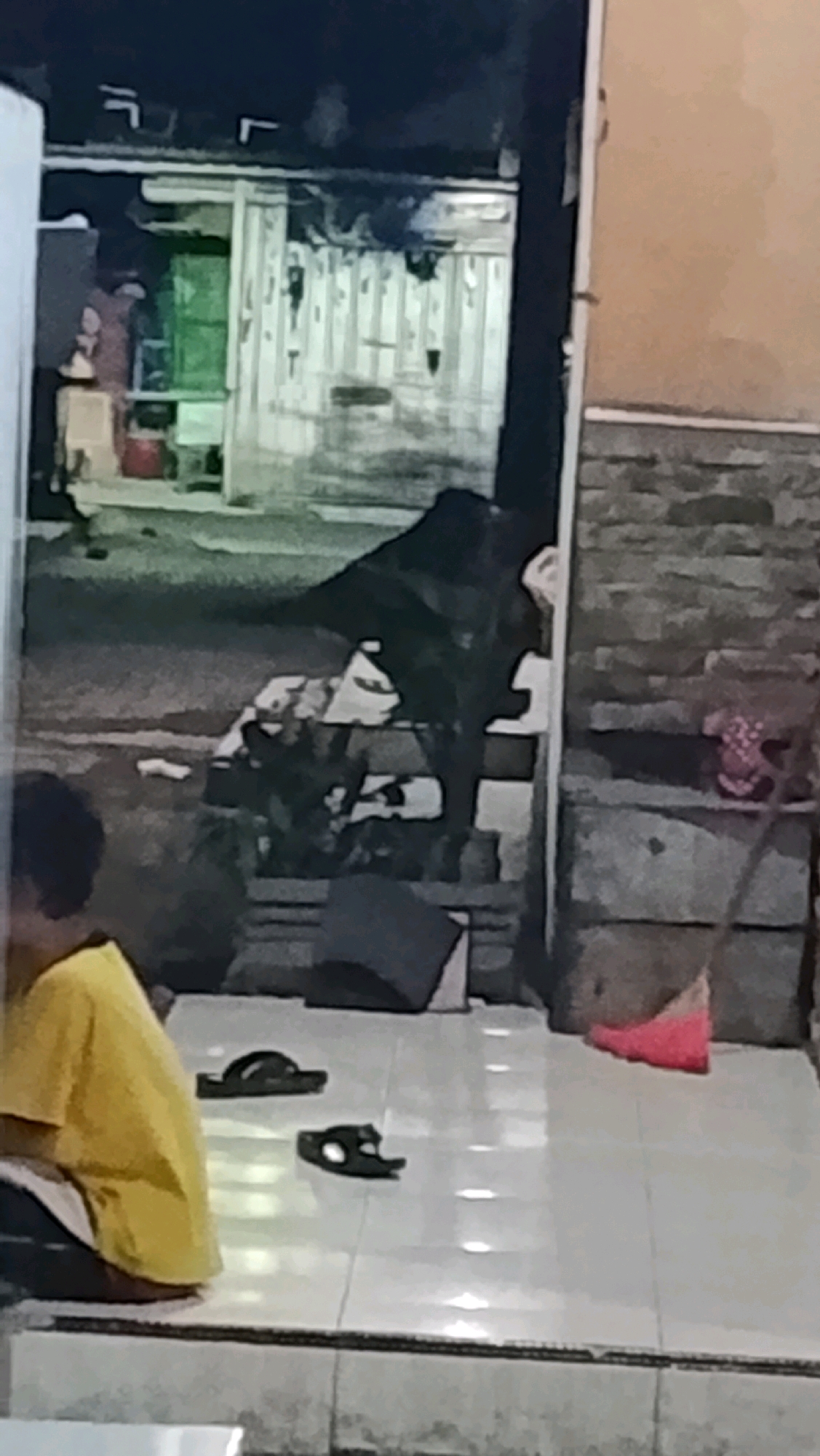 The image depicts an outdoor scene in Ujungrusi, Indonesia, likely at night or in low light conditions, given the general darkness and limited visibility. In the foreground, on a tiled floor, are a pair of dark sandals. To the left, a person wearing a yellow shirt and white pants is partially visible, seated on the floor. The background shows a building with a corrugated metal exterior. A dark, amorphous shape, possibly a large animal or a pile of dark objects, is situated in front of the building. To the right, a broom with a pink dustpan is leaning against a textured wall, suggestive of a dwelling. The overall impression is one of a simple residential area, possibly a backyard or alleyway, with a few everyday objects present.