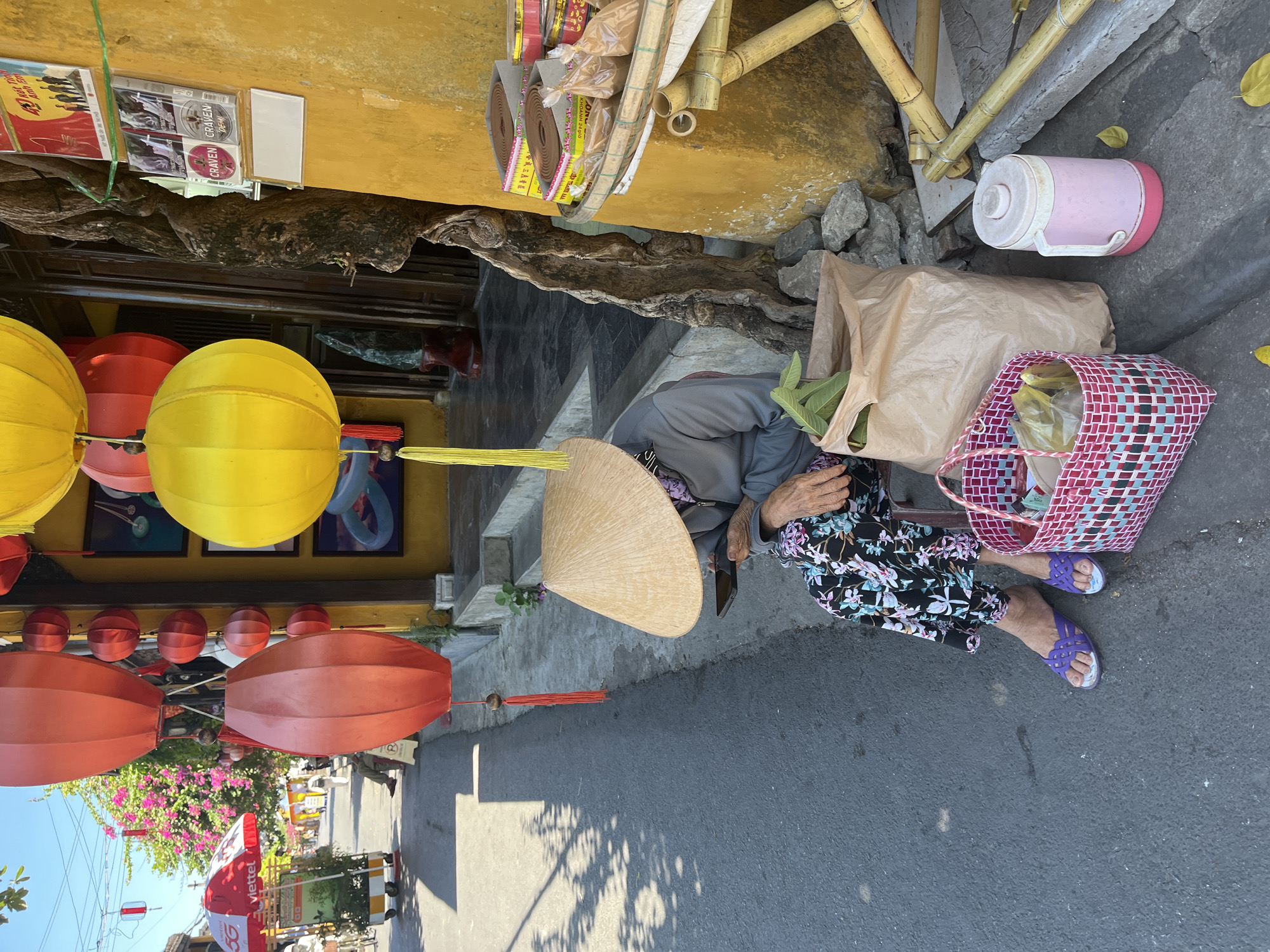 The image depicts a vibrant street scene in Hội An Ward, Vietnam, with a person resting beside a building. An elderly individual, likely a woman, is seated on a low step, wearing a grey top, a black floral skirt, and purple sandals. Her head is covered by a traditional light brown conical hat (nón lá), obscuring her face.
Next to her are a large brown paper bag with green leaves protruding, a red and white woven plastic bag containing other items, and a pink and white thermos bottle. The immediate surroundings feature a bright yellow building wall adorned with prominent tree roots. Above these, merchandise such as books or CDs (one with a red cover and text "40 NĂM CƠN BÃO CUỘC ĐỜI," others mentioning "CRAVEN") and incense coils (labeled "KHOANH 24HO") are displayed.
Numerous colorful lanterns, including large yellow and smaller red ones, hang overhead, characteristic of Hội An. The scene is illuminated by bright sunlight, casting distinct shadows on the asphalt road and concrete sidewalk. In the background, an umbrella with the "Viettel 5G" logo, flowering pink bougainvillea bushes, and other street elements are visible. The time of day is clearly sunny daylight. The person appears to be resting or waiting.