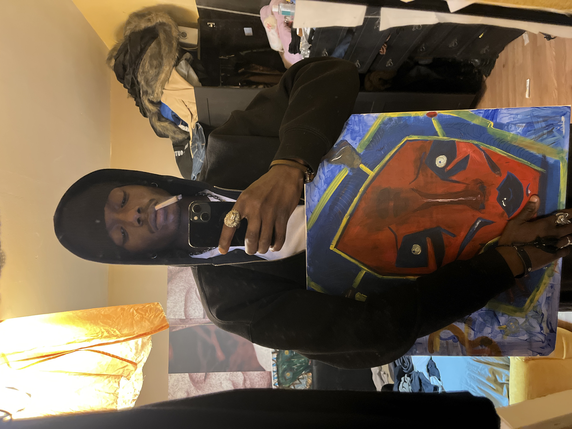 A photograph shows a dark-skinned man in a black hooded sweatshirt holding a painting. He also has a cigarette in his mouth and is holding a cell phone up to the mirror to take the image. He is wearing several rings. The painting is of a face in shades of blue, yellow, and red. It's unknown whether this image was taken in Barcelona, Spain.