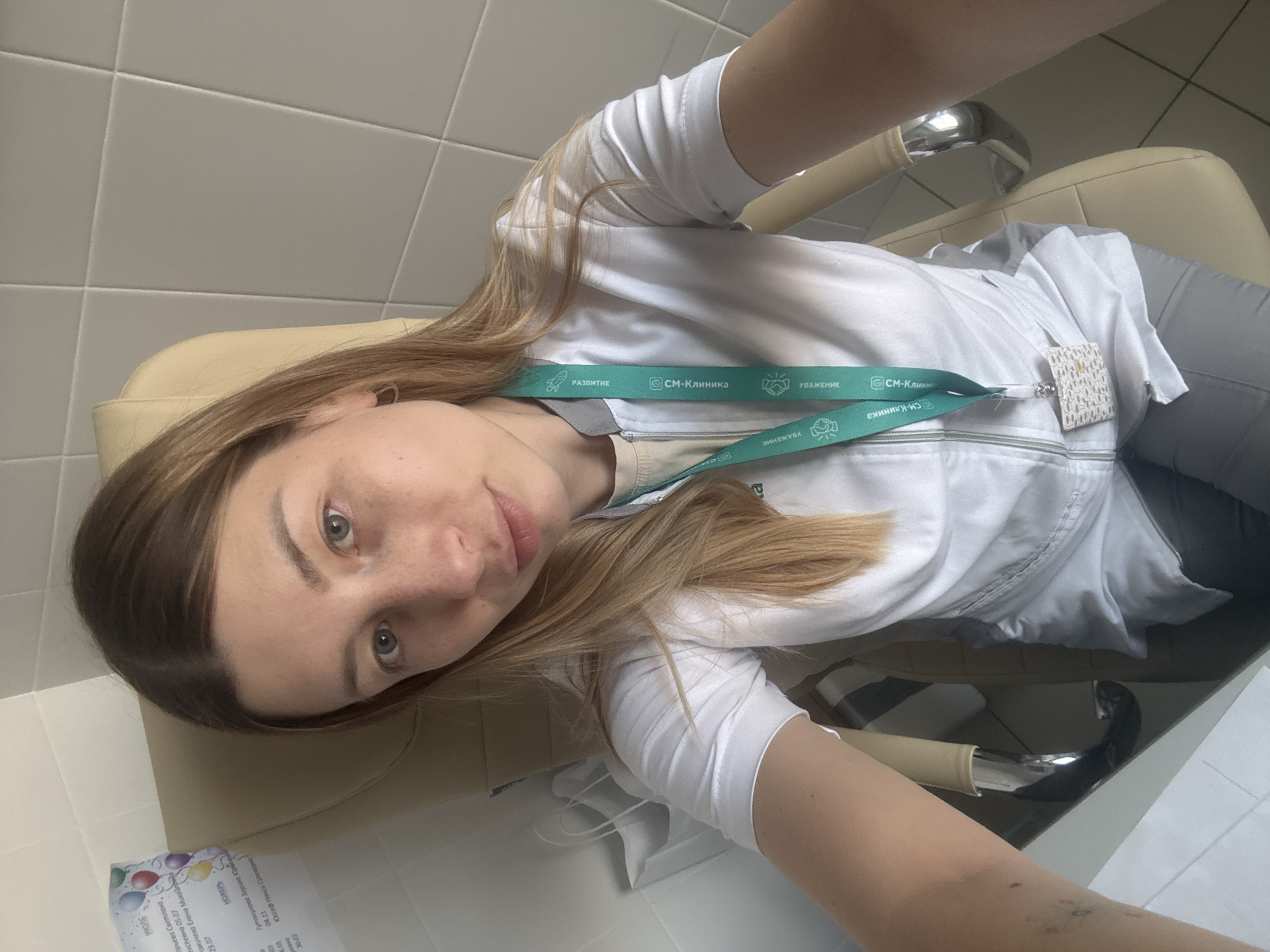 The image shows a selfie of a person wearing a white medical coat and a lanyard from "CM-Clinic". The lanyard has the words "DEVELOPMENT" and "RESPECT" written in Russian. The person is sitting in a beige chair, likely in a medical examination room. The background includes tiled walls. A paper with text in Russian, including names such as "Гутякулова Зарина Юроблена" and "Юссеф Нанси Суеплевна", as well as dates, is partially visible at the bottom left. "CM-Clinic" is located in the city of Moscow, Russia.