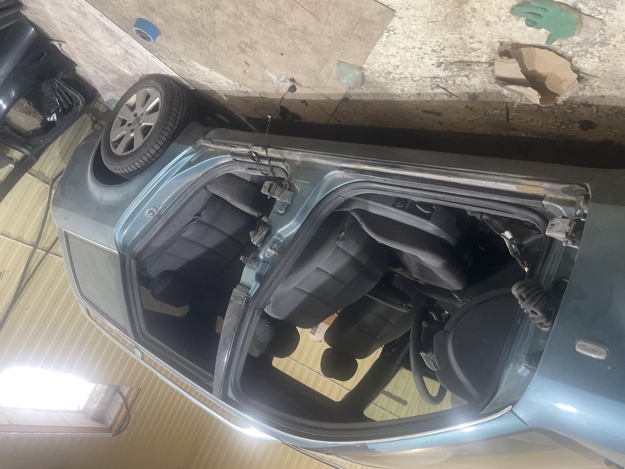 The image depicts an interior view of a teal-colored station wagon, likely in a state of repair or disassembly, situated within a garage or workshop. The driver's side door is open, revealing the interior. Visible elements include the steering wheel, dashboard, front and rear seats, and door panels. The car appears to be a right-hand drive model, indicated by the placement of the steering wheel. The floor of the garage is concrete, with scattered debris including a green work glove, a piece of cardboard, and blue painter's tape. A separate car door, possibly removed from the vehicle, is leaning against the wall on the right side of the image. The background consists of corrugated metal walls and a bright light source from above. The overall impression is of a functional, albeit messy, automotive workspace. The location is specified as Навадворскі сельскі Савет, Belarus.