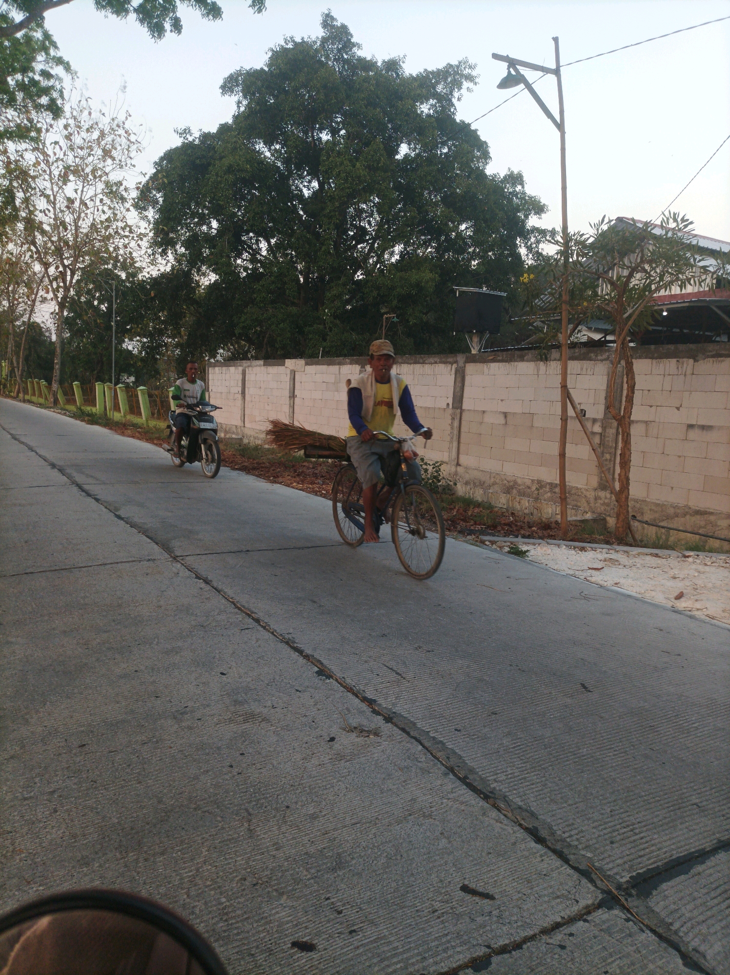 In CANGA'AN, Indonesia, two individuals are observed on a paved road. The first is a person riding a motorcycle, facing away from the viewer, on the left side of the road. The second is a man on a bicycle, facing the viewer, to the right of the motorcycle rider. He is carrying a bundle of dry brush or straw on the back of his bicycle. The road is flanked by a concrete wall on the right, with trees and vegetation visible in the background. A street lamp is also present on the right side of the road. The sky is clear and appears to be during the day. The lower left corner of the image shows a blurred reflection, possibly from a camera lens or a vehicle mirror.