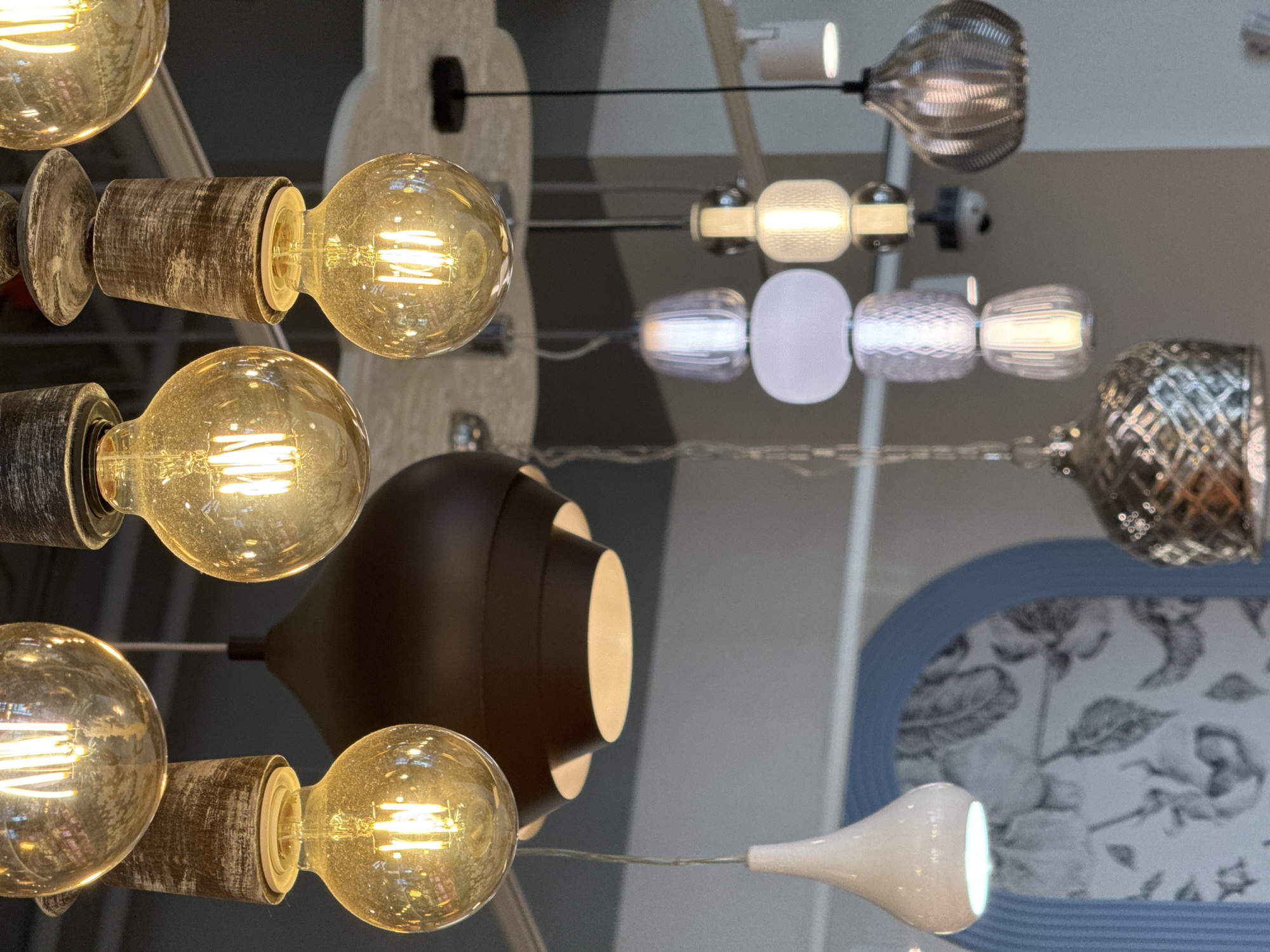 The image displays a collection of various light fixtures. A cluster of round, vintage-style bulbs with weathered, metallic casings is prominently featured on the left side. A dark brown, dome-shaped lamp with internal lighting is positioned near the center. In the background, there are decorative glass pendant lights with different textures and shapes, hung on thin wires. A patterned floral design with a blue border is visible in the lower right corner. The exact location within Moscow, Russia cannot be determined from the image.