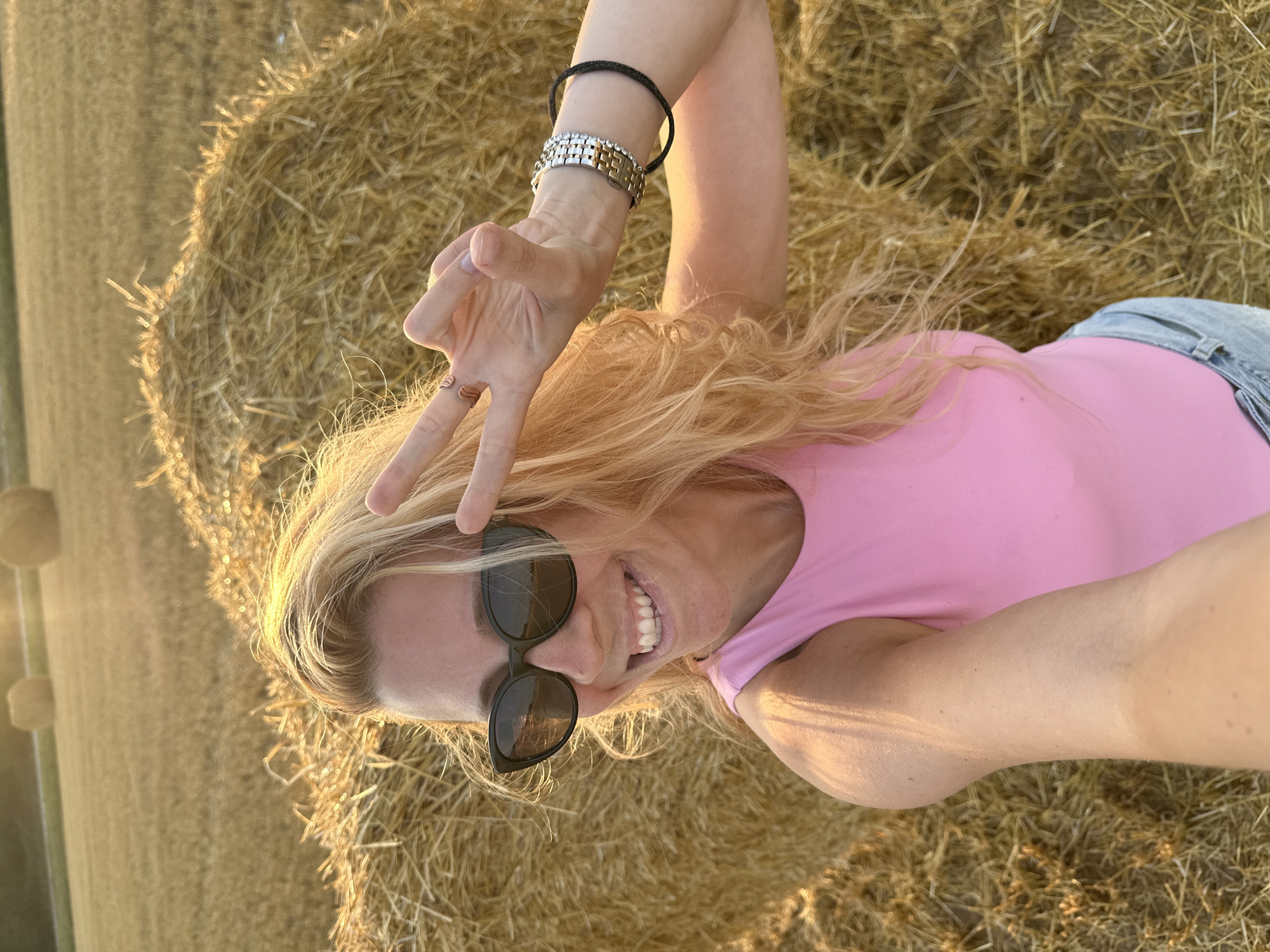 A young woman with blonde hair and sunglasses is taking a selfie in a field of hay bales. She is wearing a pink top and denim shorts. The sun is setting, casting a warm glow on the scene. The woman is smiling and holding up two fingers in a peace sign. The background shows several large hay bales and a distant hill. The location is identified as Trouville-Alliquerville, France.