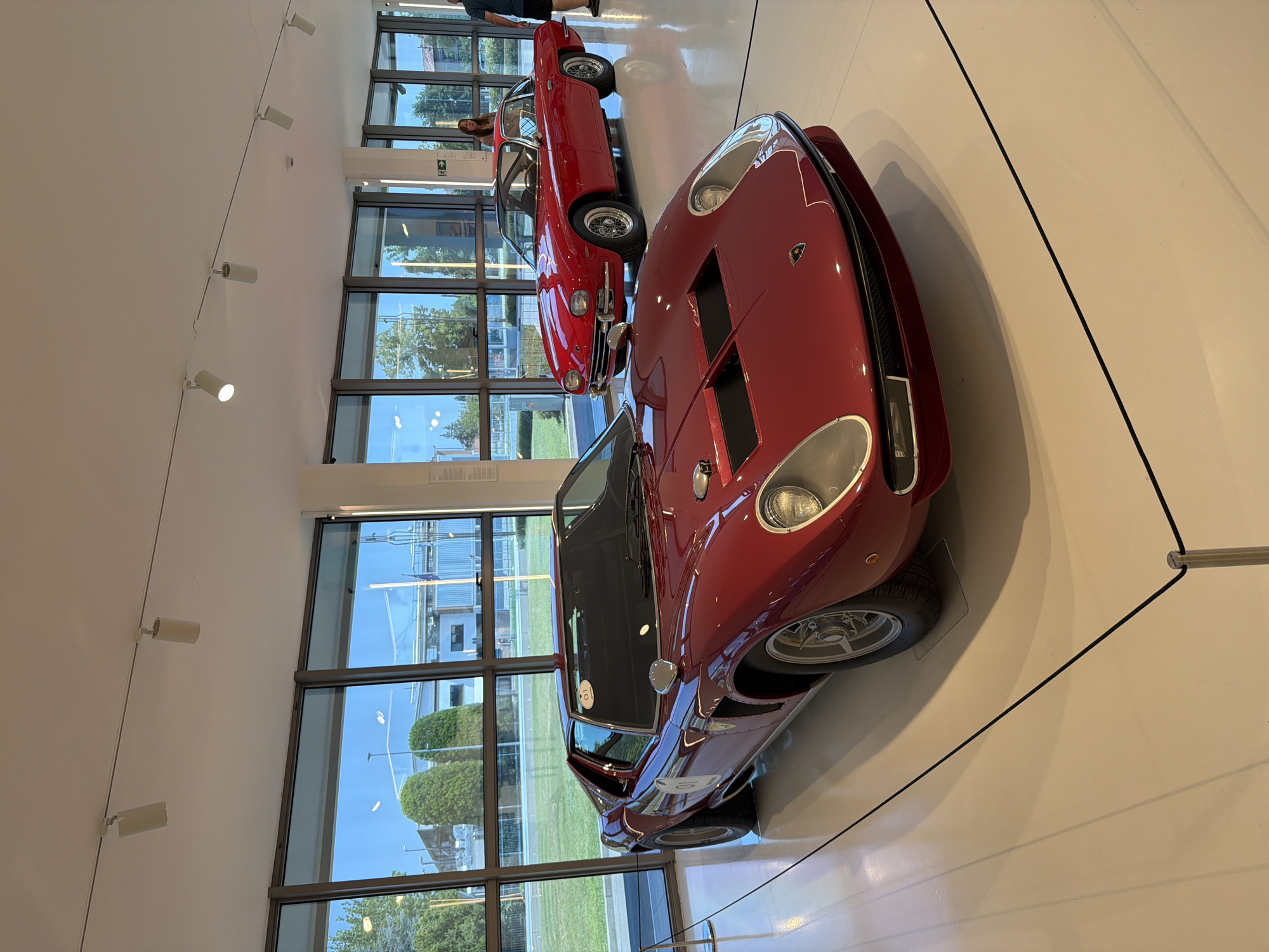 The image shows a red Lamborghini Miura parked in a well-lit showroom with large windows. In the background, another red classic car, likely a Lamborghini 400 GT 2+2, is parked. A woman is standing near the second car, looking towards the camera. The showroom floor is white and clean, with a black rope barrier in the foreground. Outside the windows, there are trees, buildings, and a blue sky. The location is the Lamborghini Museum in Sant'Agata Bolognese, Italy, which is in the province of Bologna, not Maggi.
