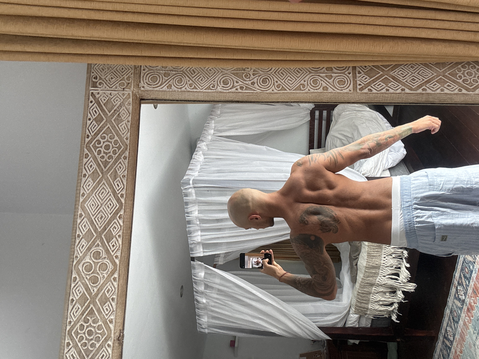 A man with a shaved head, visible tattoos on his arms and back, and wearing light blue striped pajama bottoms and white underwear, stands in a bedroom and takes a selfie in a large mirror. He is holding up a smartphone in his right hand, pointing it towards the mirror. The bedroom features a large bed with a dark wood frame, draped with white sheer curtains. A macrame-style bedspread with fringe is draped over the foot of the bed. The mirror frame is intricately carved with geometric and circular patterns. To the right of the mirror, a floor-to-ceiling golden curtain hangs. The flooring visible in the foreground is a patterned rug over dark wood. The scene suggests a domestic setting, likely in a hotel room or residence in Kuta Selatan, Indonesia, given the tropical ambiance suggested by the decor and curtains.