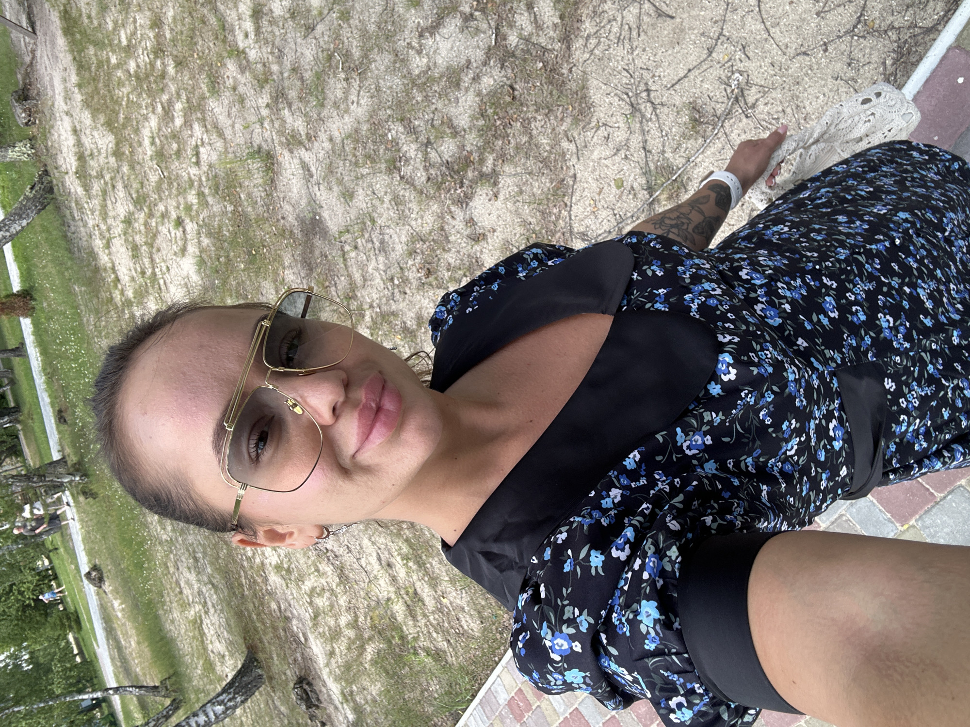 The image shows a person taking a selfie outdoors. The person is wearing large, gold-rimmed sunglasses and a black dress with a blue floral pattern. They have a tattoo on their left arm. In the background, there is a sandy area with sparse vegetation, and in the distance, there are trees and a path where other people can be seen walking. The lighting suggests it is daytime, and the weather appears to be fair. The person's expression is neutral, and they are holding a white, lacy object in their right hand. The scene appears to be a park or a natural area. There is no visible text in the image.