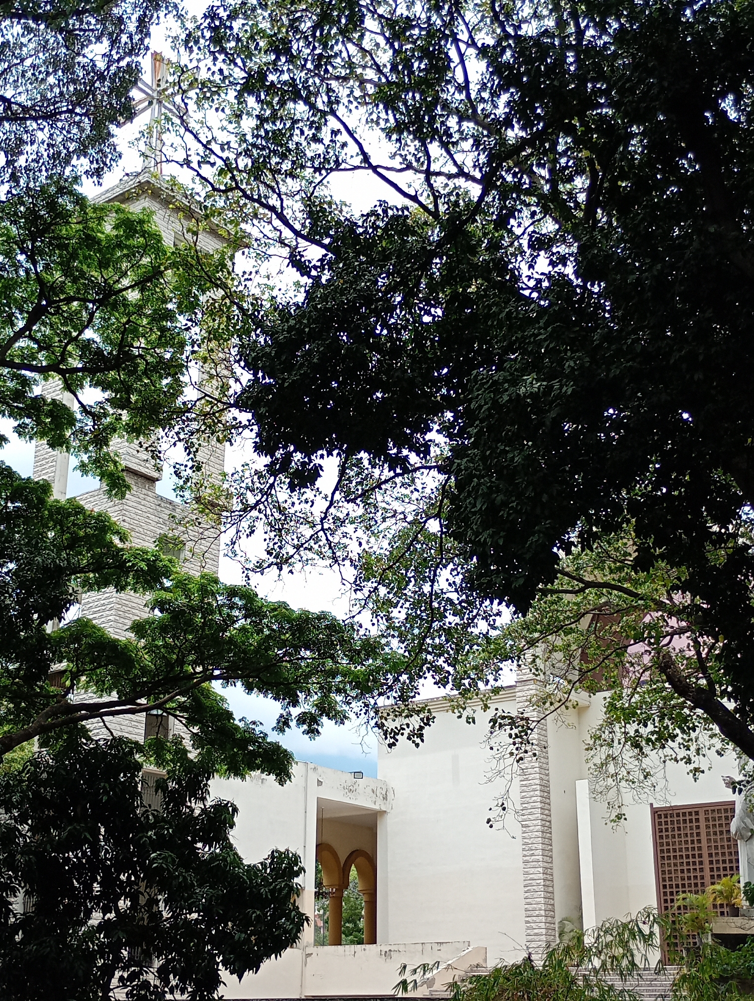 The image shows a white building with a tower and cross on top, partially obscured by trees. The building appears to be a church or religious institution, with architectural details like arches and a latticed window visible. The vegetation suggests a tropical or subtropical environment. The photo's perspective is from a low angle, looking up at the building. The location is the Chapel of the Catholic University Andres Bello (UCAB) in Caracas, Venezuela.