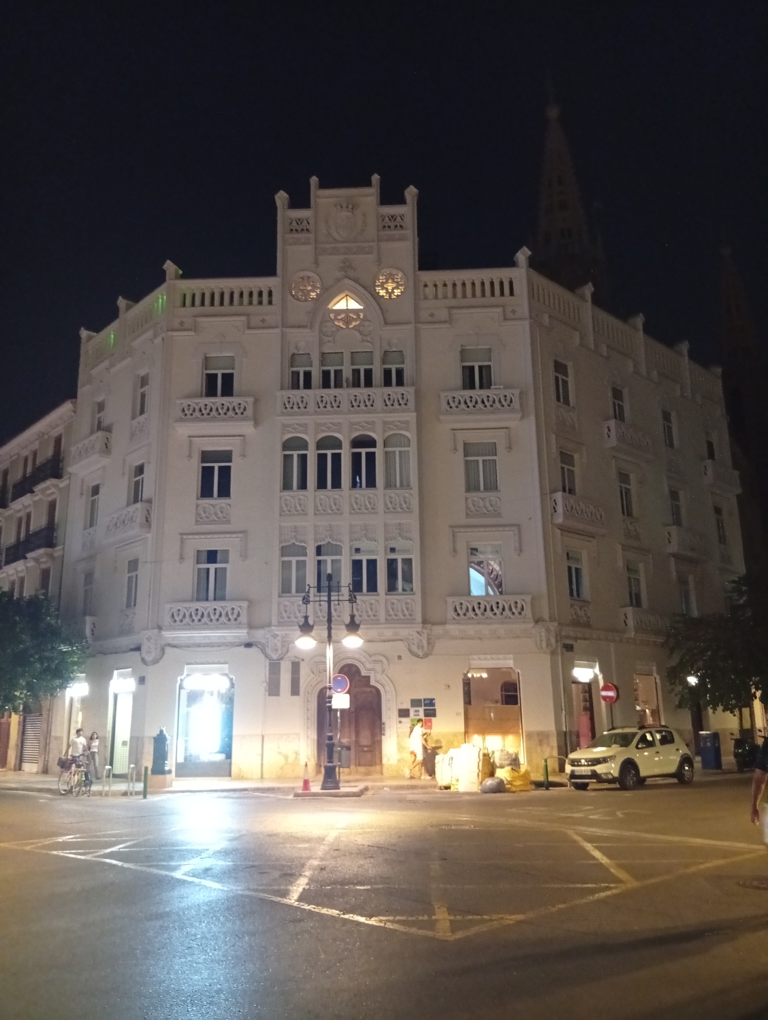 The image depicts a street scene at night in Valencia, Spain, likely on a corner where two streets intersect, as indicated by the road markings. A large, ornate, cream-colored building with multiple floors dominates the frame. The building features several decorative balconies with intricate ironwork, arched windows, and a distinct ornamental facade on its upper levels. A tall, pointed spire, possibly part of a church or cathedral, is visible in the background against the dark night sky. Streetlights illuminate the scene, casting light on the wet asphalt road and the building's facade.
In the foreground, a white car is parked on the right side of the street. On the left side, two individuals are standing near bicycles and a lamppost. One individual appears to be a woman, and the other a man, both standing and looking in the direction of the building. The street is wet, suggesting recent rain, and reflects the light from the lamps and the building. Several trash bins are also visible along the sidewalk. The overall atmosphere is quiet and urban, illuminated by artificial light.