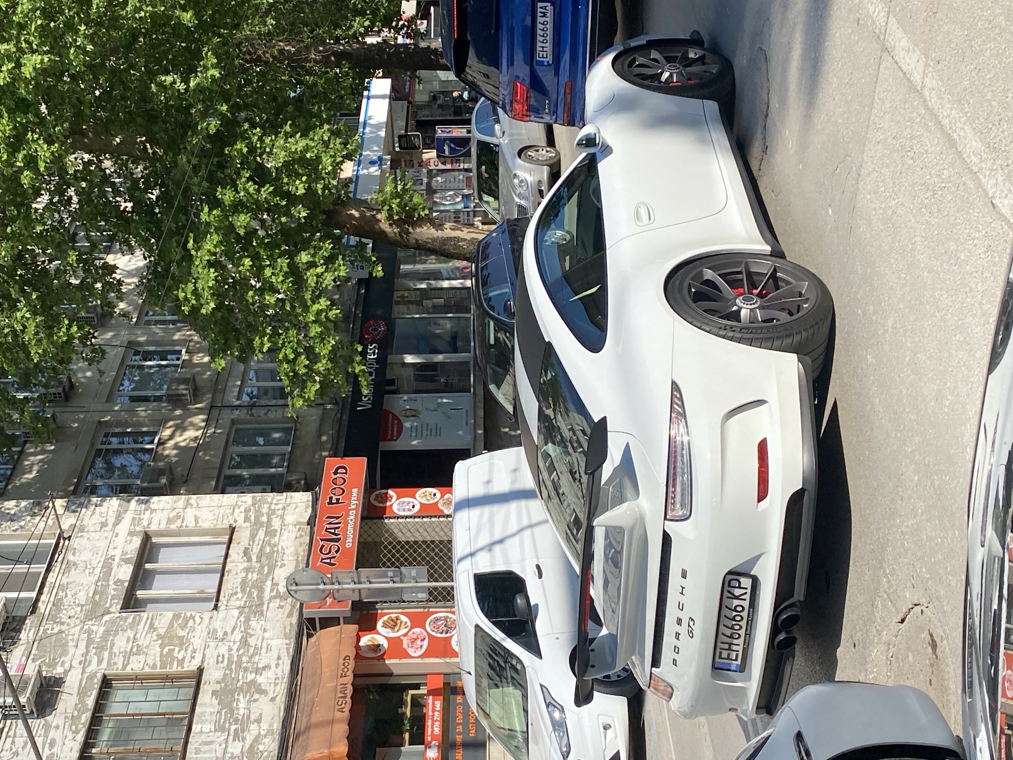 The image depicts a street scene in Pleven, Bulgaria. A white Porsche GT3 is parked on the street. The car's license plate is EH 6666 KP and has the marking "Porsche Zentrum Kassel". Behind the Porsche is a white van. The building next to the vehicles has two businesses: "Asian Food" and "Vision Express". Another car behind the Porsche has the license plate "EH 6666 MA". There is a large tree visible on the left side of the frame.