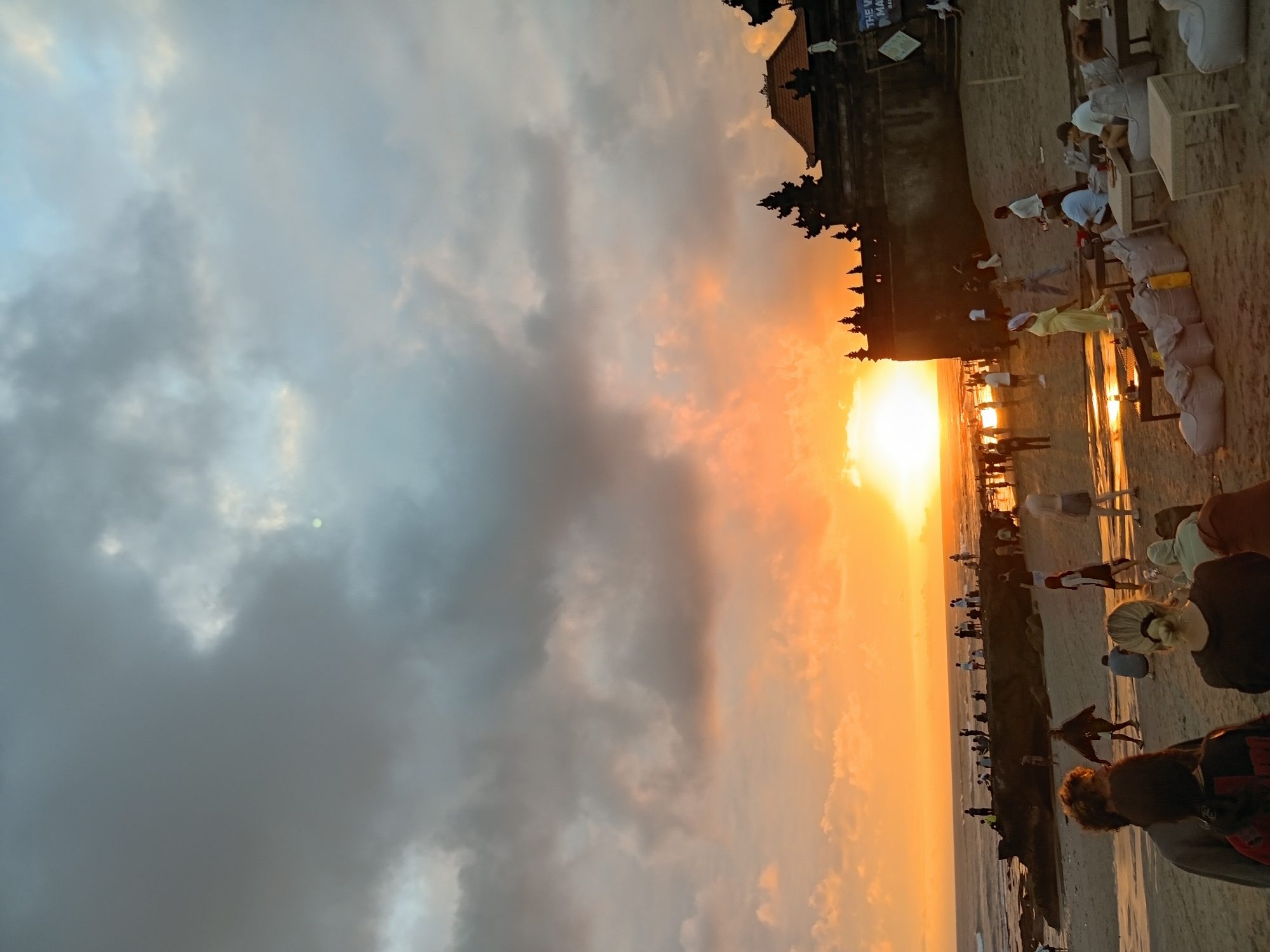 In North Kuta, Indonesia, a beach scene unfolds at sunset. The sky is dominated by a dramatic display of clouds, with warm orange and yellow hues radiating from the setting sun. The sun itself is a bright orb on the horizon, casting a golden glow over the ocean and the wet sand.
A group of people are visible on the beach. Some are gathered near the water's edge, while others are spread out on the sand. In the foreground, several individuals are seated at low tables with beanbag chairs, appearing to be relaxing or socializing. Further back, a crowd of people are observing the sunset, some standing and some sitting. A few people are also seen walking along the beach and in the water.
On the right side of the image, a traditional Balinese temple structure is silhouetted against the sky. The temple has distinct tiered roofs and ornate carvings. The beach itself is sandy, with some rocky outcrops near the water. The waves are gently rolling onto the shore. The overall atmosphere is serene and picturesque, capturing a moment of natural beauty and human activity at dusk.