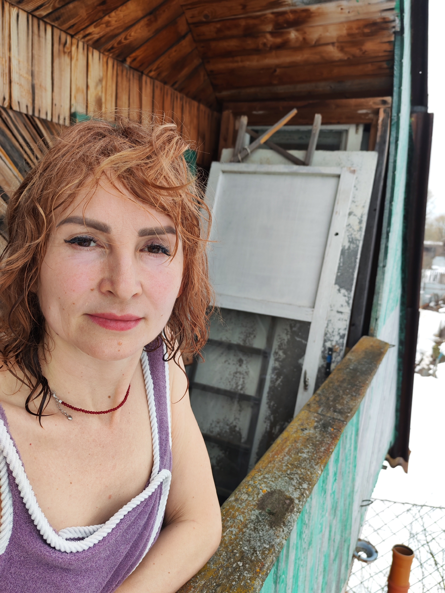 A woman with wet, reddish-brown hair stands on a balcony. She wears a purple top with white trim and a red beaded necklace. The balcony has a weathered green railing. Behind her are stacked white-framed windows and what appears to be a wooden tool leaning against them. The balcony is attached to a wooden structure with weathered brown planks. The background shows a snowy landscape with glimpses of buildings. The image was taken in Chaikovsky, Russia.