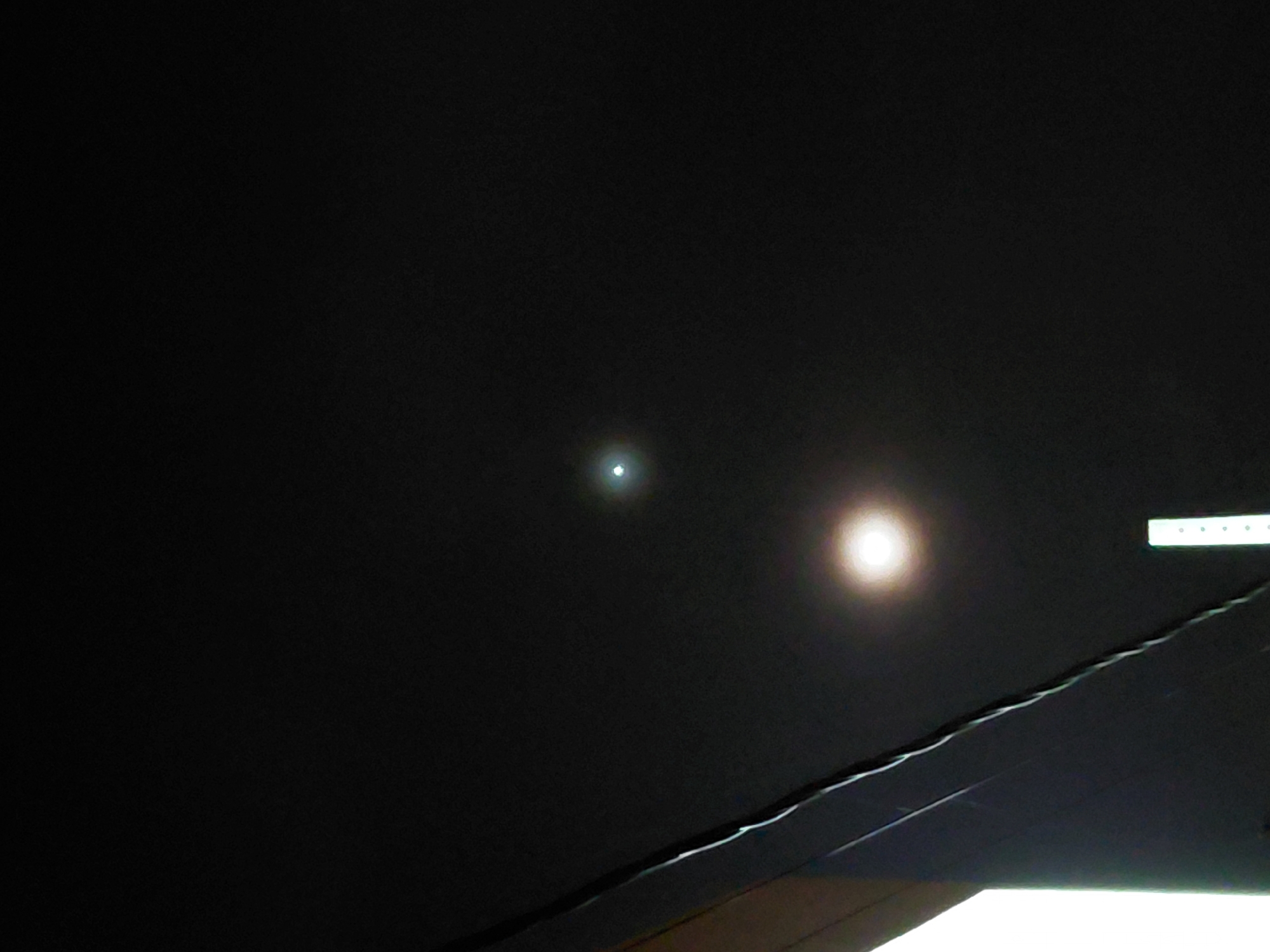 The image shows a dark, night sky over Encarnacion, Paraguay, with two distinct light sources. One is a bright, diffuse light near the bottom center of the frame, likely an artificial light source such as a streetlamp or building light, casting a yellowish glow. Above and to the left of this, a smaller, more concentrated point of light is visible, appearing as a bright white orb, possibly the moon or a distant star. A diagonal dark object, possibly a building edge or a power line, occupies the lower left portion of the frame, obscuring a sliver of a brightly lit area. A thin, vertical object with a series of small white dots is also visible in the lower center of the image. The overall scene is dark and appears to be an outdoor urban or semi-urban environment.
