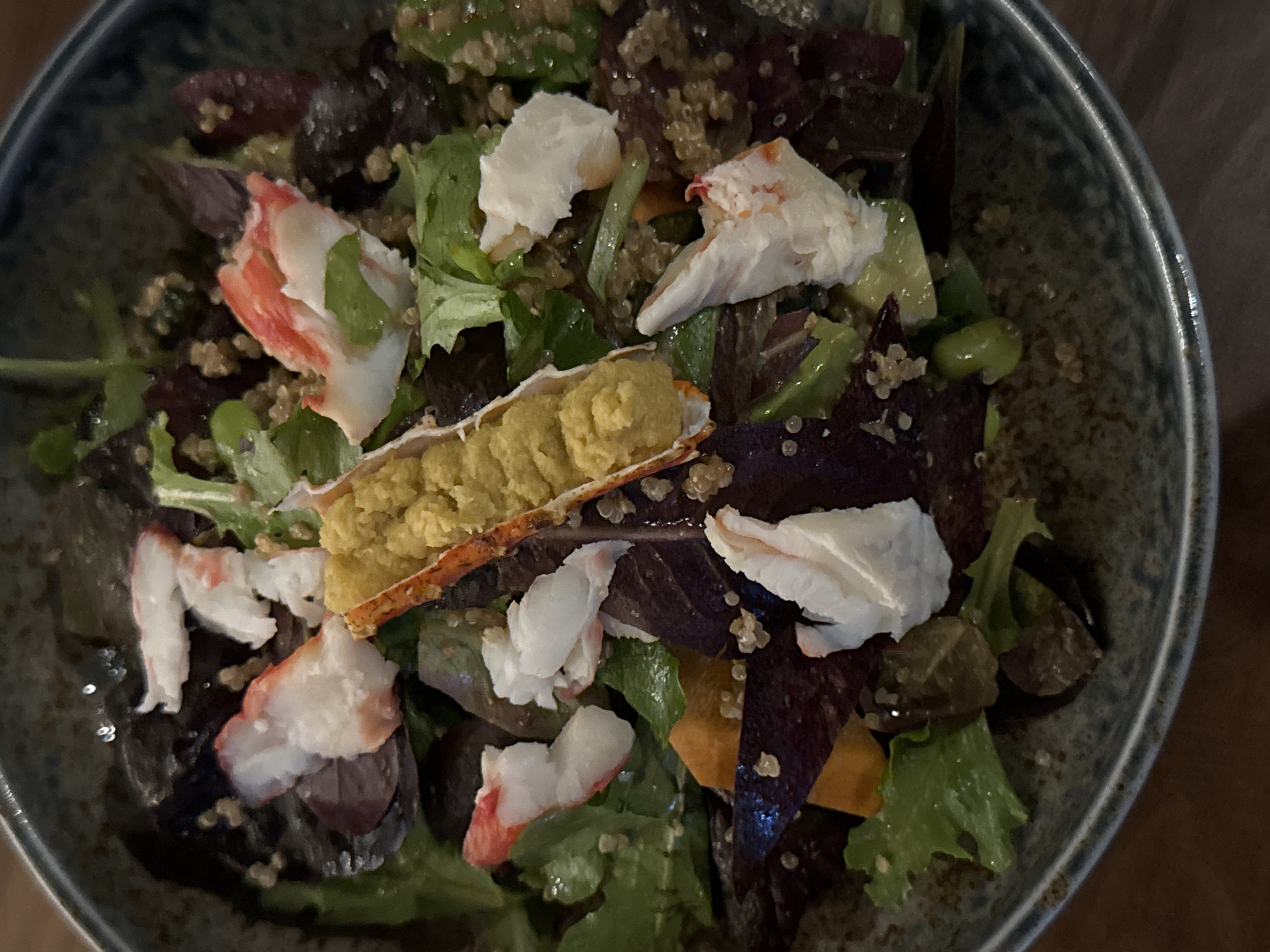 A close-up, overhead shot shows a salad in a dark, textured bowl. The salad contains chunks of white and pink seafood, possibly lobster or crab, which are scattered throughout. There are also dark red beet slices, green leafy vegetables, edamame beans, and quinoa. A small piece of orange carrot or sweet potato is visible. The background is blurred and appears to be a wooden surface. The lighting is dim, creating a moody atmosphere. The image does not contain any identifiable landmarks or clues indicating a specific location in Sofia, Bulgaria.