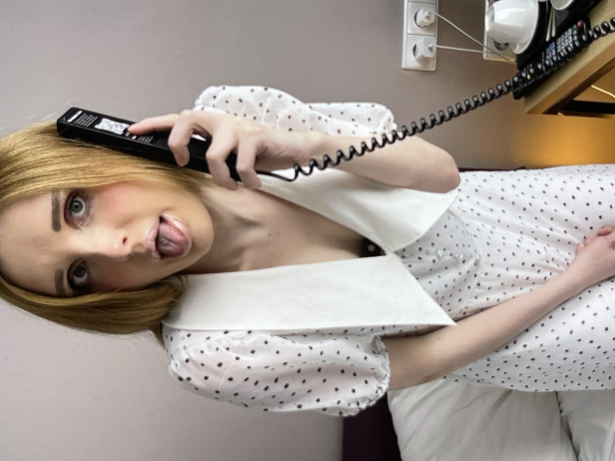 A young woman with blonde hair is holding a black telephone receiver to her right ear and her tongue is sticking out. She is wearing a white dress with black polka dots and a large white collar. She is standing in what appears to be a hotel room, with a bed on the left and a desk with a telephone and electrical outlets on the right. The wall is a light pink color.