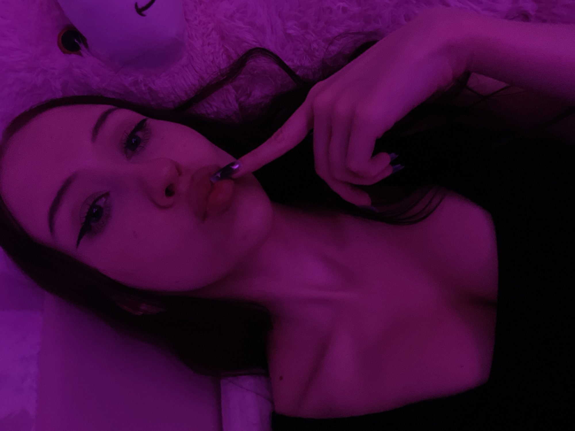 The image shows a close-up, overhead shot of a young woman with dark hair, wearing a black top. She is lying down, with her face turned towards the camera and her finger placed on her lips, in a "shushing" gesture. The scene is lit with pink and purple light, casting a strong hue over everything. A plush, light-colored object, possibly a stuffed animal, is visible to her right. The background appears to be a soft, textured surface. There is no indication of the location being in Votkinsk, Russia.