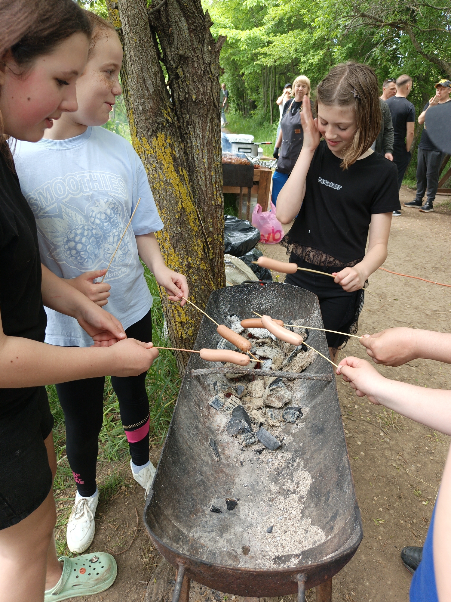 In Терновский округ, Ukraine, a group of people are gathered around a makeshift barbecue grill, possibly constructed from a modified wheelbarrow. Several individuals are skewering sausages on wooden sticks and roasting them over charcoal embers. The barbecue is positioned on a dirt path, with a tree nearby. Other people are milling about in the background, some near tables with food and drinks. The attire of the individuals varies, from casual t-shirts and shorts to leggings and sneakers. The setting suggests a casual outdoor gathering, possibly a picnic or informal barbecue.