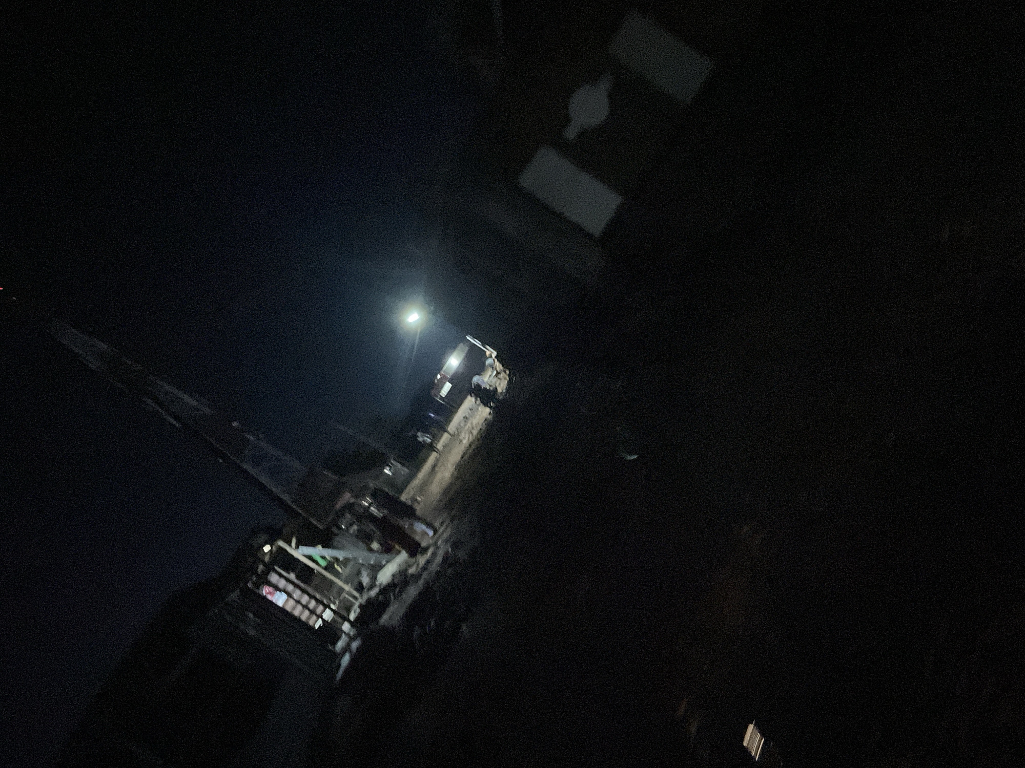 In the dark, a partially constructed building stands in Lokoja, Nigeria. A ladder is visible inside the building. Several individuals are seen near the building and a vehicle. The ground appears to be dirt. The scene is lit by artificial light sources.