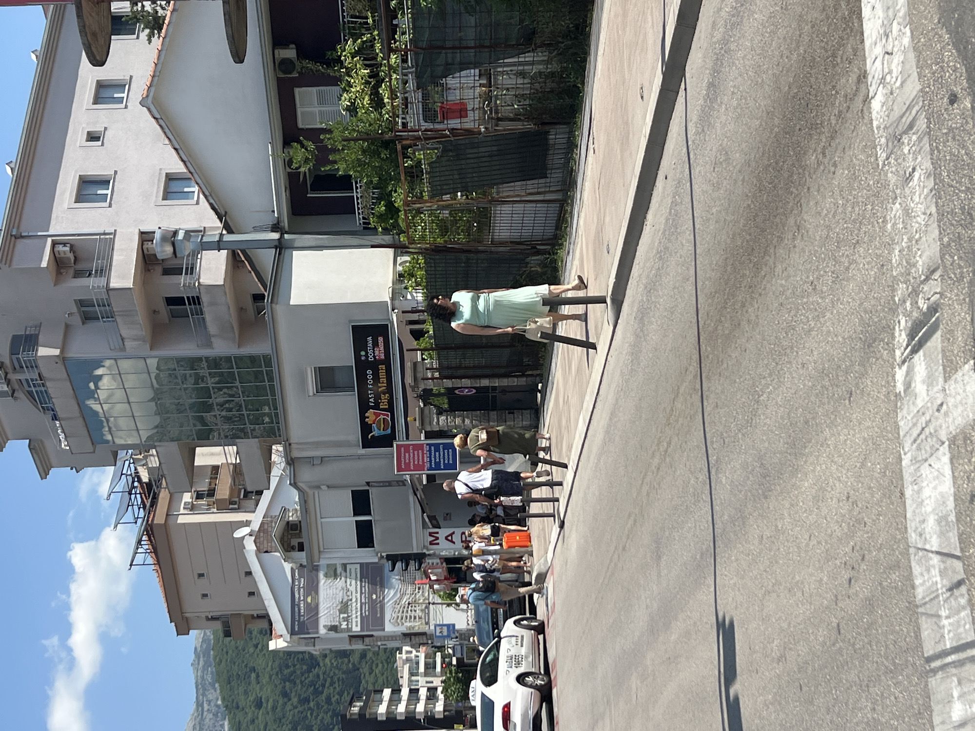 The image shows a street scene in Budva, Montenegro, on a sunny day. In the foreground, a grey asphalt road with a white pedestrian crossing is visible. On the right side of the road, a woman in a light green dress walks on the sidewalk, holding a beige handbag. She is walking away from the camera. The sidewalk is bordered by a metal fence and greenery.
Further down the street, a group of people are standing on the sidewalk, some with luggage, suggesting they might be waiting for transportation. An orange suitcase is prominent among them. A white taxi with the number 19655 is parked on the left side of the road, facing away from the camera. Behind the taxi, a black car is also present.
Several buildings line the street. On the right, a building with a dark red facade and white shutters is visible, with green vegetation growing on balconies. To the left of this, a building with a glass facade reflects the sky and clouds. Further left, there are more buildings, some appearing to be multi-story residential or commercial properties. A sign for "FAST FOOD BIG MAMA" is visible on one of the buildings. Mountains covered in green trees form the backdrop of the city. Traffic lights are also visible, indicating an intersection. The sky is blue with scattered white clouds.