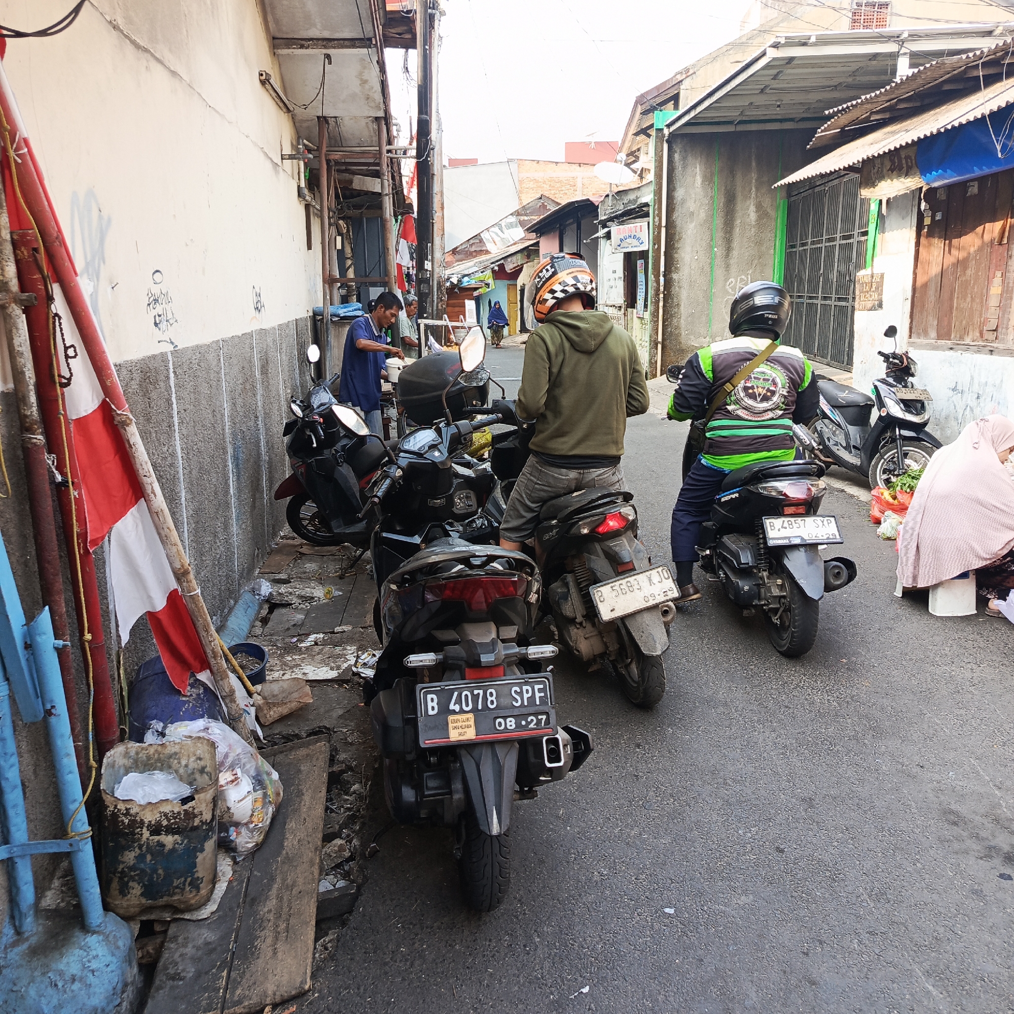 In a narrow urban alleyway in South Jakarta, Indonesia, three motorcycles are parked. The foreground features a red and white Indonesian flag tied to a bamboo pole. To the left, a man in a blue shirt is serving food from a cart, with a second man standing nearby. Further down the alley, two men on motorcycles are visible, one wearing a green hoodie and the other a black and green jacket. Buildings line both sides of the alley, some with corrugated metal roofs and others with exposed brickwork. The ground is paved with asphalt and shows signs of wear and debris. A woman in a light pink hijab is seated near a stall on the right side of the alley, beside a pile of fresh produce. License plates on the motorcycles indicate they are registered in Jakarta. The scene appears to be a bustling street food vendor area.