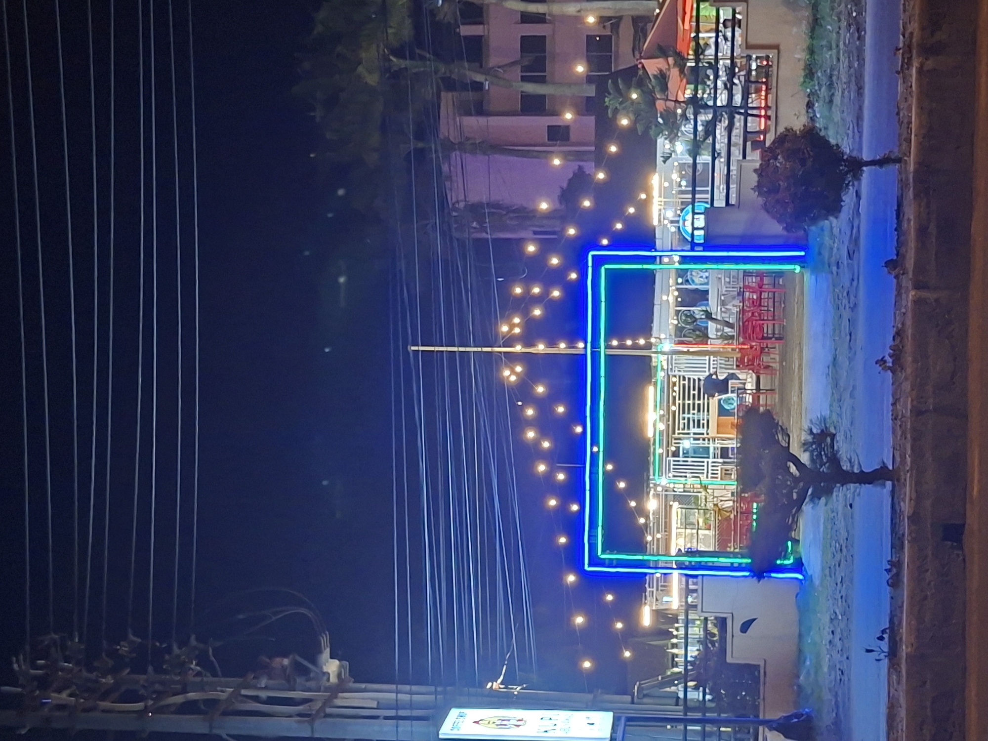 The image shows a nighttime view of a commercial establishment in Lhokseumawe, Indonesia, with a facade brightly lit by blue and green neon. Several lines stretch across the sky. String lights decorate the building's perimeter and interior. Some trees flank the building. A few tables and chairs can be seen through the windows. There is another tall building in the background. A small white illuminated sign is in the foreground. The image is shot from ground level, looking slightly upwards.