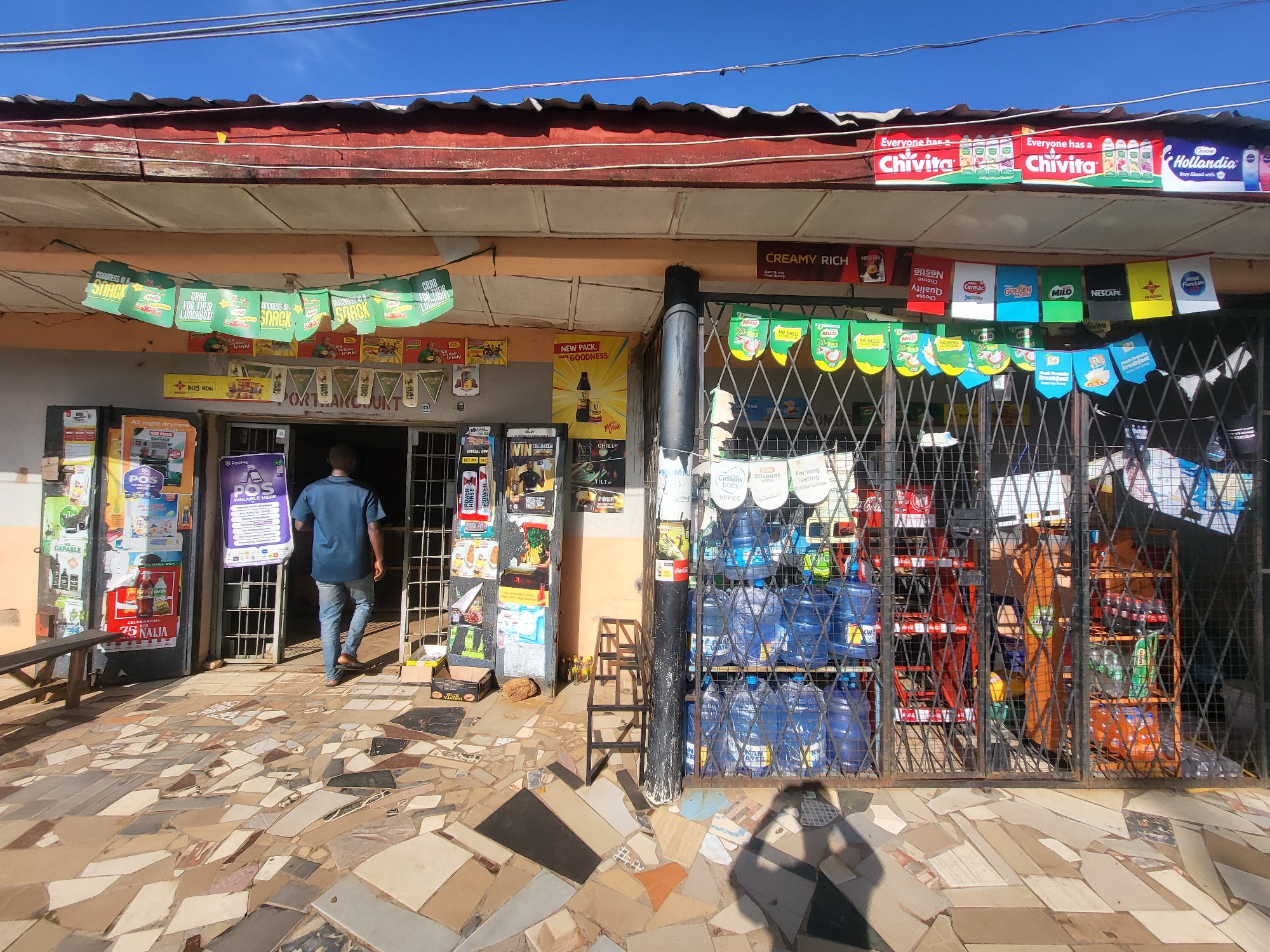 The image depicts a street-level view of a small shop in Barakin Akawo, Nigeria. The shop has a red-tiled roof and is decorated with numerous colorful banners advertising various products, including "Chivita" and "Hollandia". Inside, shelves are stocked with water jugs, bottled drinks, and other retail items. A man in a blue shirt and jeans is walking into the shop. The ground in front of the store is paved with irregularly shaped tiles in various shades of brown, grey, and beige. The scene is brightly lit by the sun, casting distinct shadows on the ground. The shop is secured with a metal grate.