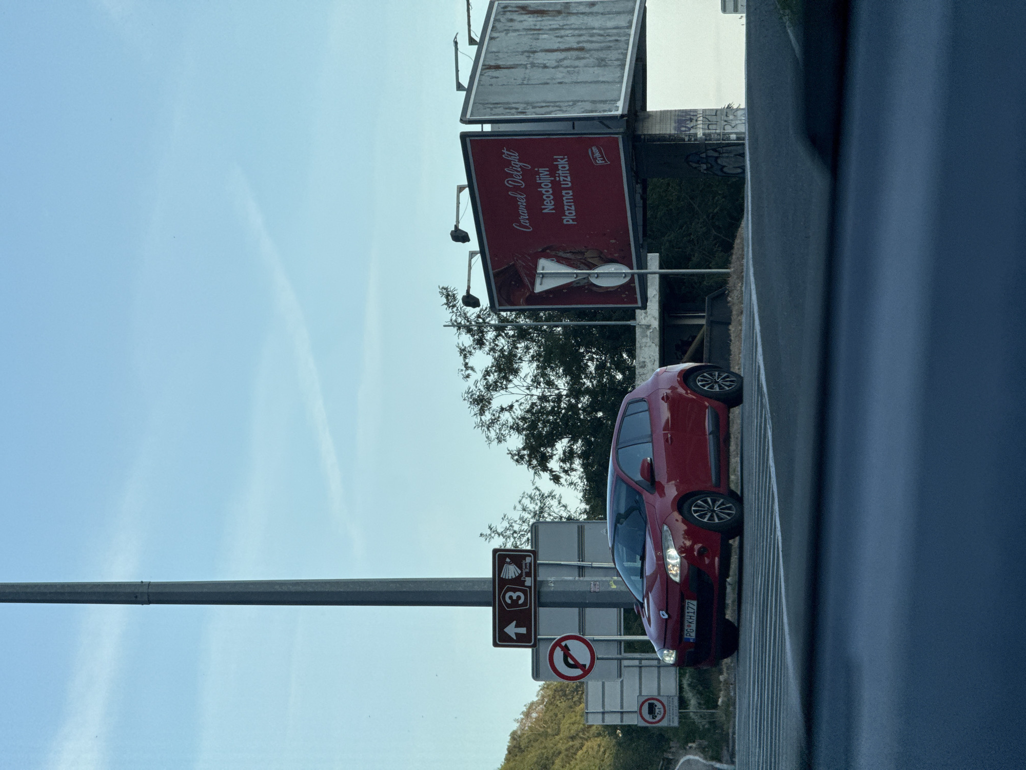 A red Renault Megane is stopped at a crosswalk on an overcast day, with a tall pole and a billboard on the right. The car has its headlights on and its license plate reads "PG KH177". The billboard has an advertisement for "Caramel Delight" with text that appears to be in a foreign language. Other signs are visible, including one with an arrow indicating directions and a no-left-turn sign. The environment is a road intersection, possibly in a city, with some vegetation in the background. The sky is clear and blue.