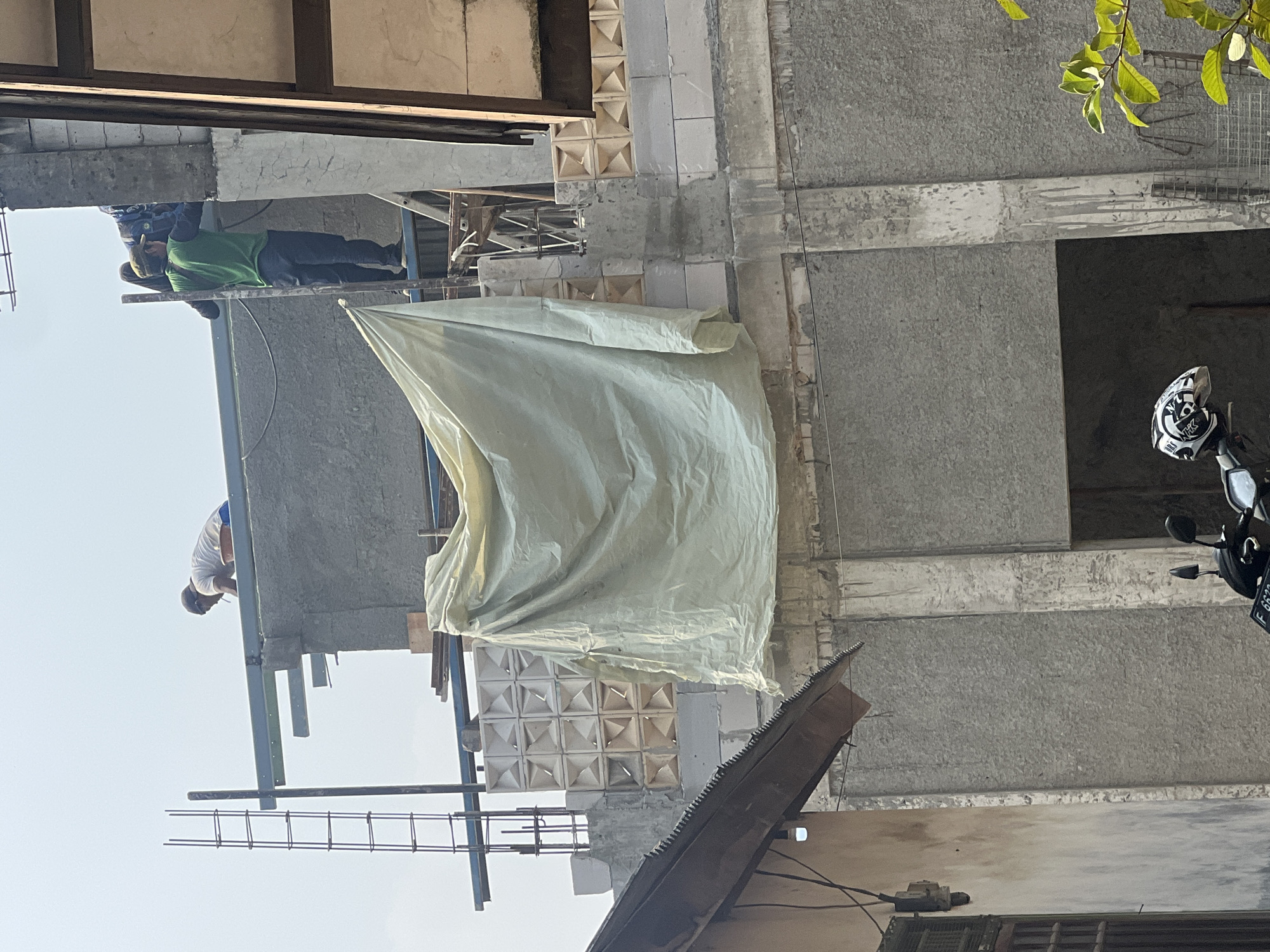 The image depicts a construction site in Klapanunggal, Indonesia, with an unfinished building under construction. Two construction workers are visible on the roof. One worker, wearing a white t-shirt and blue shorts, is bent over near the edge of the roof. Another worker, dressed in a green long-sleeved shirt and dark pants, stands further back on the roof, facing away from the camera. Construction materials, including metal beams and rebar, are present. A large, light-colored tarp hangs from the side of the building, covering a section of the upper floor. Below the construction area, a portion of a house with a corrugated metal roof and an electrical meter is visible. A motorcycle with a helmet on its seat is parked in the foreground. The sky is clear and bright, suggesting a sunny day.