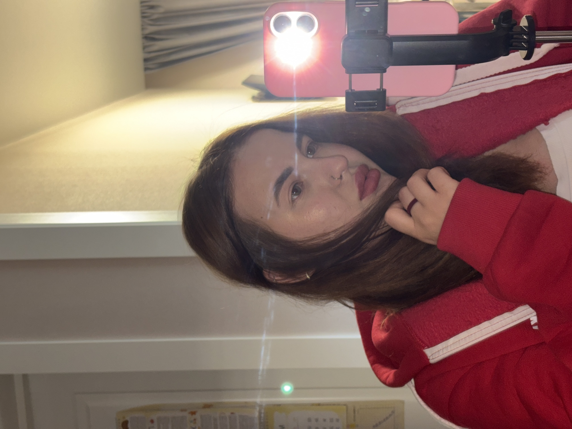 The image displays a head-and-shoulders portrait of a young woman, captured indoors in what is likely Krasnodar, Russia. The photograph is oriented 90 degrees clockwise from vertical. The woman is positioned centrally, gazing directly into the camera with a neutral to slightly posed expression. Her medium-brown hair frames her face, and her right hand is gently placed near her hair on the left side of the frame (as the image is currently rotated). She is wearing a red hooded sweatshirt featuring white trim, with a glimpse of a white garment underneath. Notable accessories include a small nose ring in her left nostril and a subtle hoop earring in her right ear.
A pink smartphone, secured in a black mounting clip (suggesting a selfie stick or stand), is prominently visible in the upper part of the frame. The phone's flash is active and brightly illuminated, serving as the primary light source and casting a strong glow that creates noticeable lens flares and reflections across the image. The background consists of light-colored indoor walls. Below the subject, a blurred lower section reveals what appears to be a shelf or desk, with faint outlines of objects such as a box and some papers or a calendar, though no specific details or legible text are discernible. The strong flash dominates the lighting, highlighting the subject while the background remains somewhat indistinct.