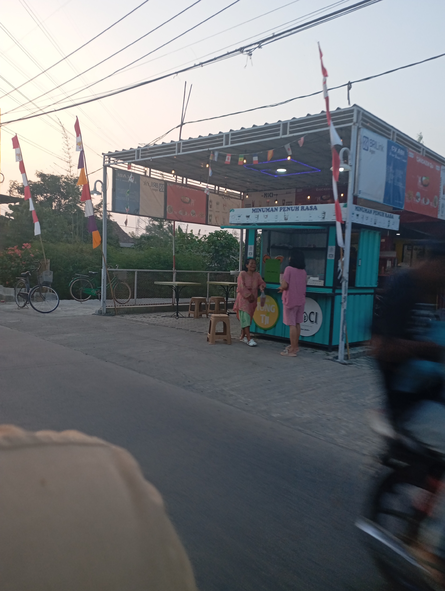 The image depicts a street scene in Kecamatan Haurgeulis, Indonesia, likely during the late afternoon or early evening given the soft, diffused light. The main focus is a small food stall, a bright teal container structure with a corrugated metal roof. The stall is adorned with Indonesian flags and various promotional banners, one reading "MINUMAN PENUH RASA" (Drinks Full of Flavor). Two women are visible near the stall; one, wearing a pink outfit, stands facing away from the camera, while another, in a peach-colored tunic and green pants, is positioned closer to the stall and appears to be interacting with someone inside or gesturing.
In the background, there are trees and a residential building with a dark roof, indicating a semi-urban or suburban setting. To the left of the stall, two bicycles are parked. The street in the foreground is asphalt, and a motorcycle, blurred due to motion, is passing by, suggesting the perspective is from another vehicle. Power lines are strung across the top of the frame, a common sight in Indonesian streetscapes. The overall atmosphere is casual and appears to be a typical commercial area.