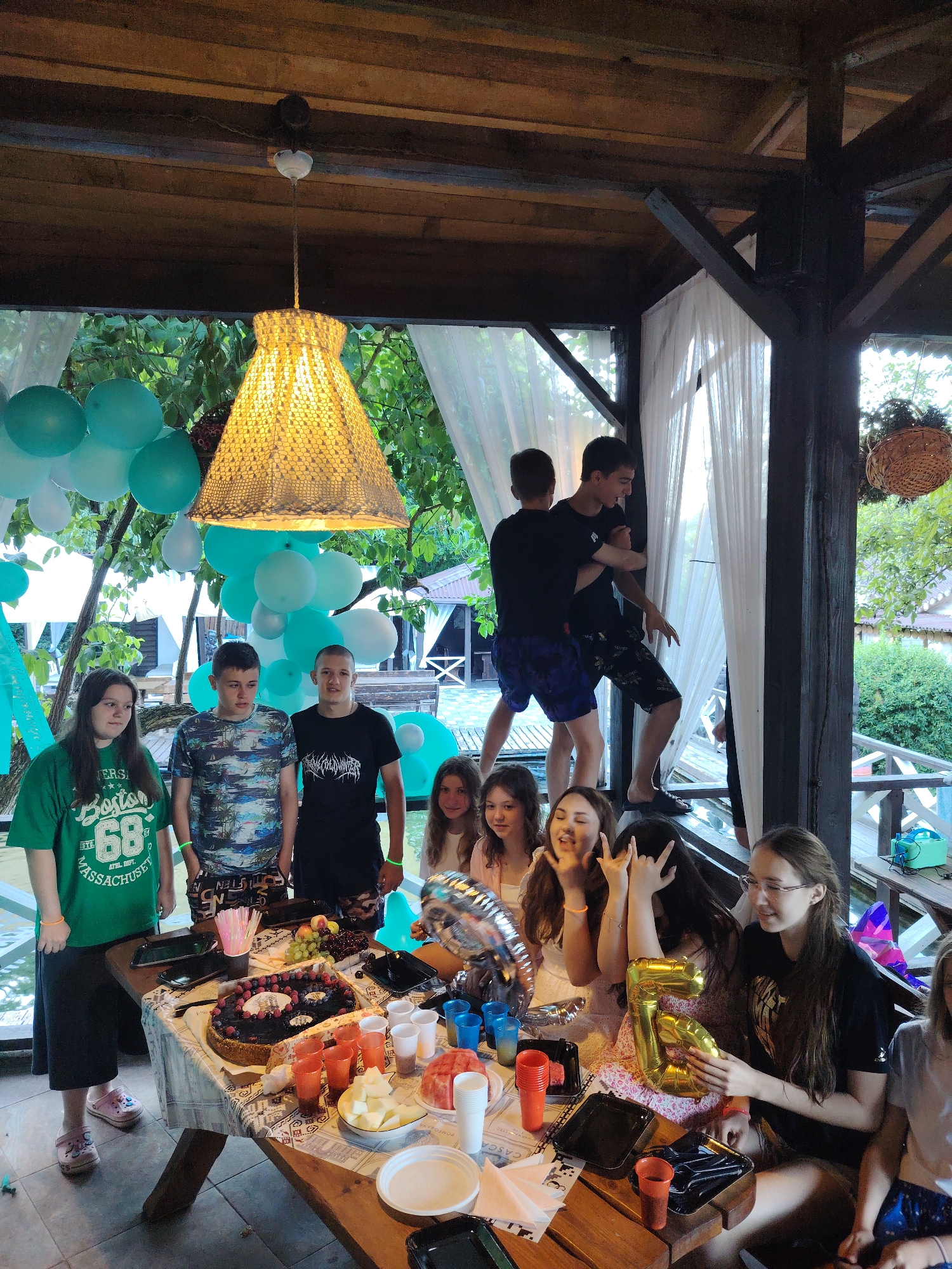 This image depicts a gathering, possibly a birthday celebration, at an outdoor covered pavilion. The pavilion has a wooden roof and support beams. White sheer curtains hang along the sides. The ground appears to be paved with tiles, and a body of water is visible behind the table. Greenery, including trees and bushes, surrounds the area, and a house with a red roof is visible in the background.
There are at least ten individuals present, a mix of young men and women. Some are standing around a table laden with food and drinks, while others are interacting further back in the pavilion. On the table, a cake with berries and candles is prominent. There are also plates of fruit, sliced fruit, cups, and a large gold balloon in the shape of the number "5". Some people are holding drinks or plates, and some are posing for the camera. Two young men in the background appear to be engaged in playful wrestling or jostling near a window or opening. The overall atmosphere seems festive and casual. The image does not provide specific information to determine its location within the city of Rostov-on-Don, Russia.