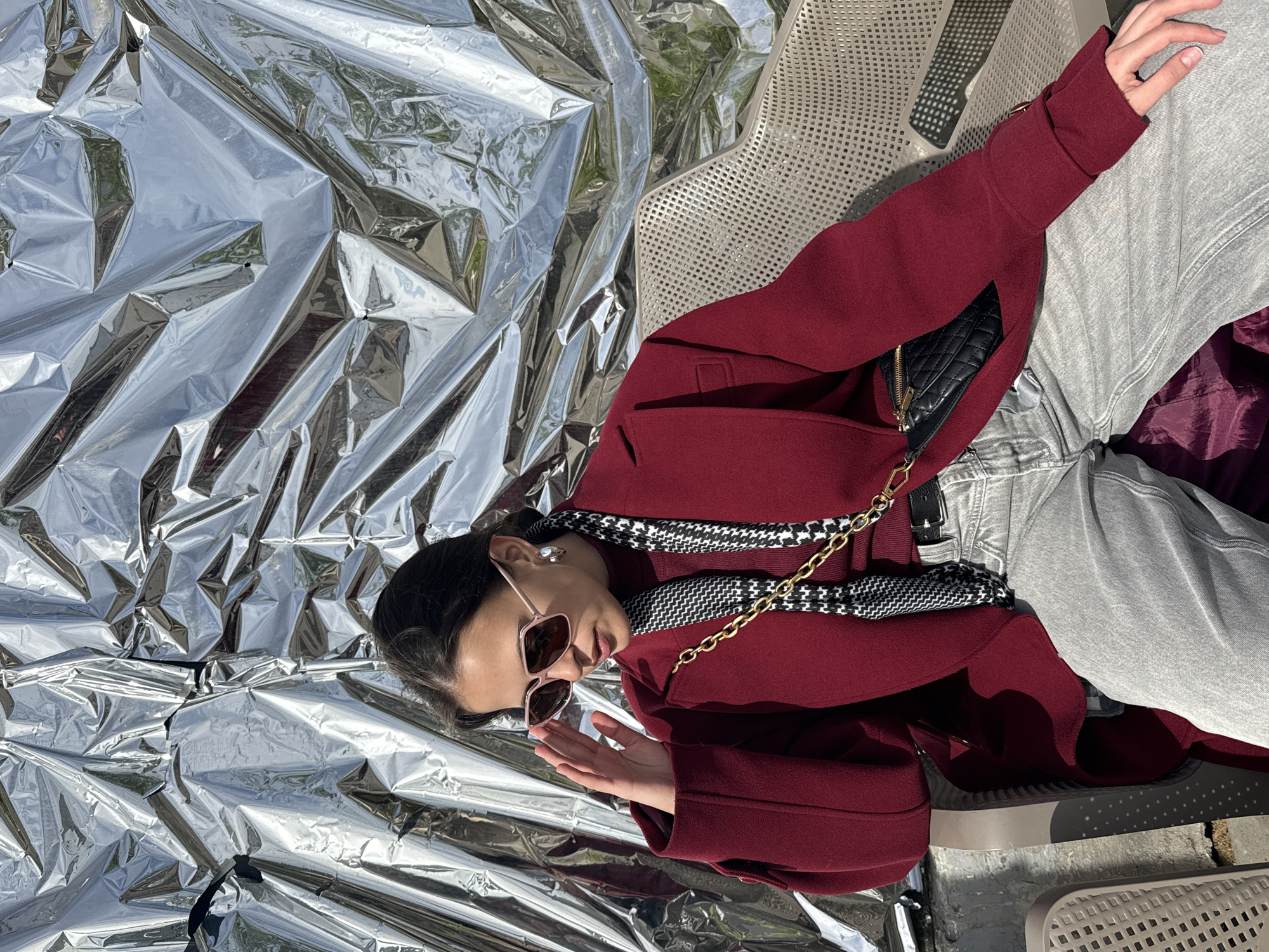 A young woman wearing sunglasses and a maroon coat lies on her back on a metallic reflective surface. Her head rests on the metallic surface, while her body is propped up by a chair. A black and white houndstooth scarf and a black quilted purse with a gold chain are draped across her coat. She wears light-wash jeans and is positioned on a plastic outdoor chair. The reflective surface dominates the background, obscuring the surroundings. The location is suggested to be in Saint Petersburg, Russia, though specific landmarks are not visible.
